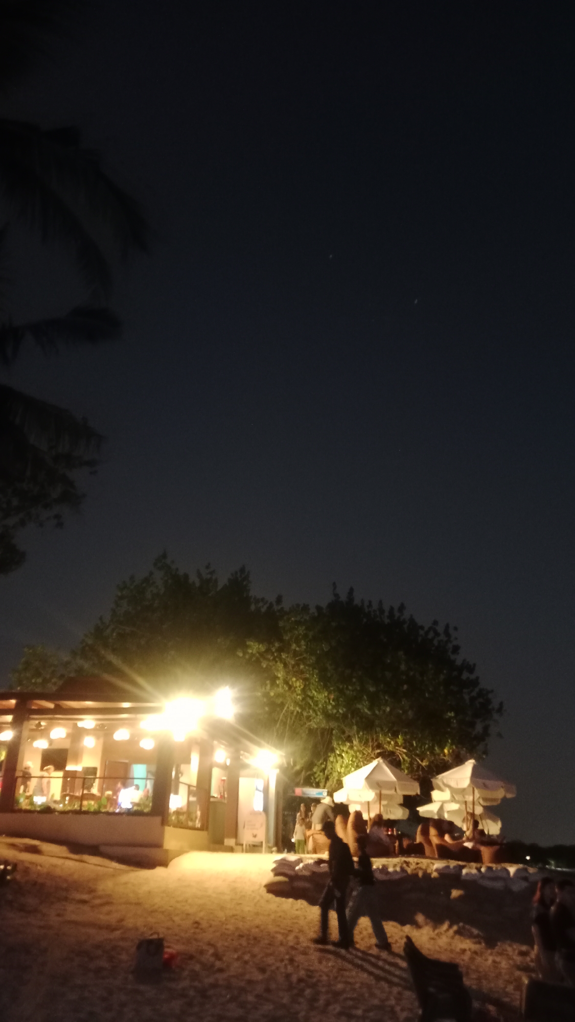 A nighttime scene depicts a beachside establishment in Kuta, Indonesia. The foreground is a sandy beach with a few scattered bags. Two individuals, silhouetted against the lights of the establishment, walk across the sand. Behind them, a structure with a thatched roof is illuminated by numerous lights, showcasing tables and chairs within, as well as people seated inside. In front of the establishment, several white beach umbrellas are set up. Lush green trees provide a backdrop, and the dark night sky is visible above, with a few faint stars or distant lights. The overall ambiance is that of a relaxed, outdoor dining or lounging area at night.