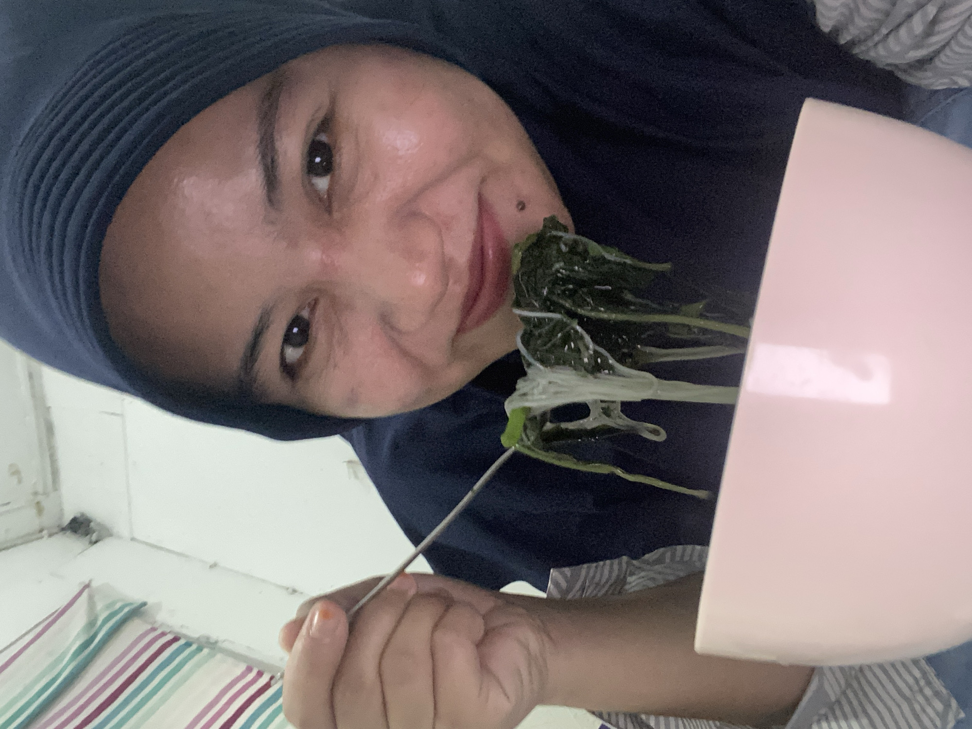 A close-up selfie shows a woman wearing a dark blue hijab and a striped shirt. She is holding a fork with noodles and green vegetables, bringing it towards her mouth. A light pink bowl is visible in the foreground. The background consists of a white wall with some horizontal stripes on the left side. The scene appears to be indoors, possibly in a residential setting. The lighting is bright, suggesting artificial light. The woman has light brown skin and is smiling slightly.