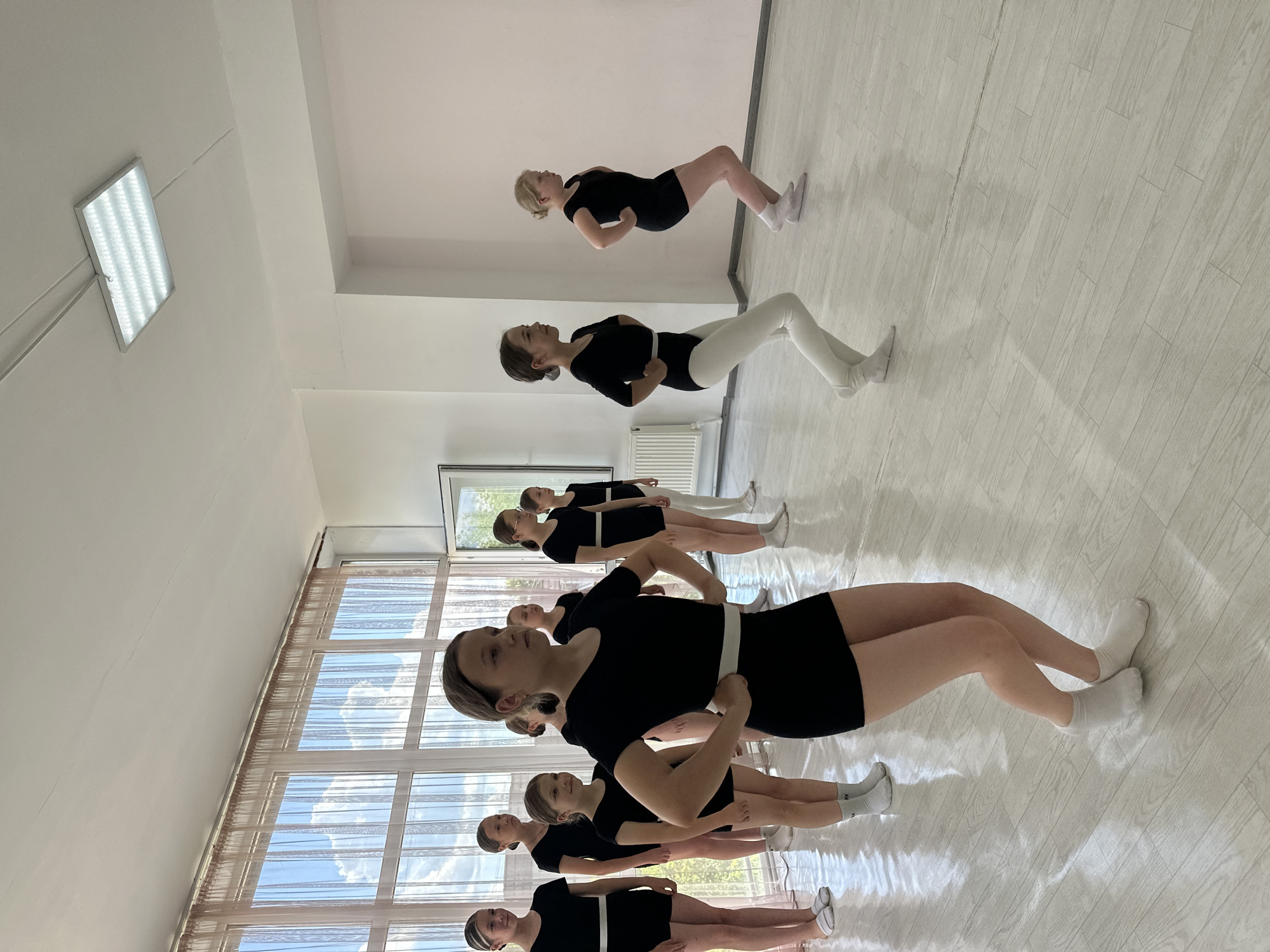 A dance class is in session inside a well-lit room. The room is located in Votkinsk, Russia. Nine females are participating in the class, wearing black leotards. Most of them have white bands around their waist. They are standing in a line. The front eight are wearing black leotards and white socks. The second woman in the line is wearing white leggings instead of black shorts. A white radiator is in the background near the window. The window has light curtains with a view of a blue sky. The floor is light-colored, possibly wood or laminate.