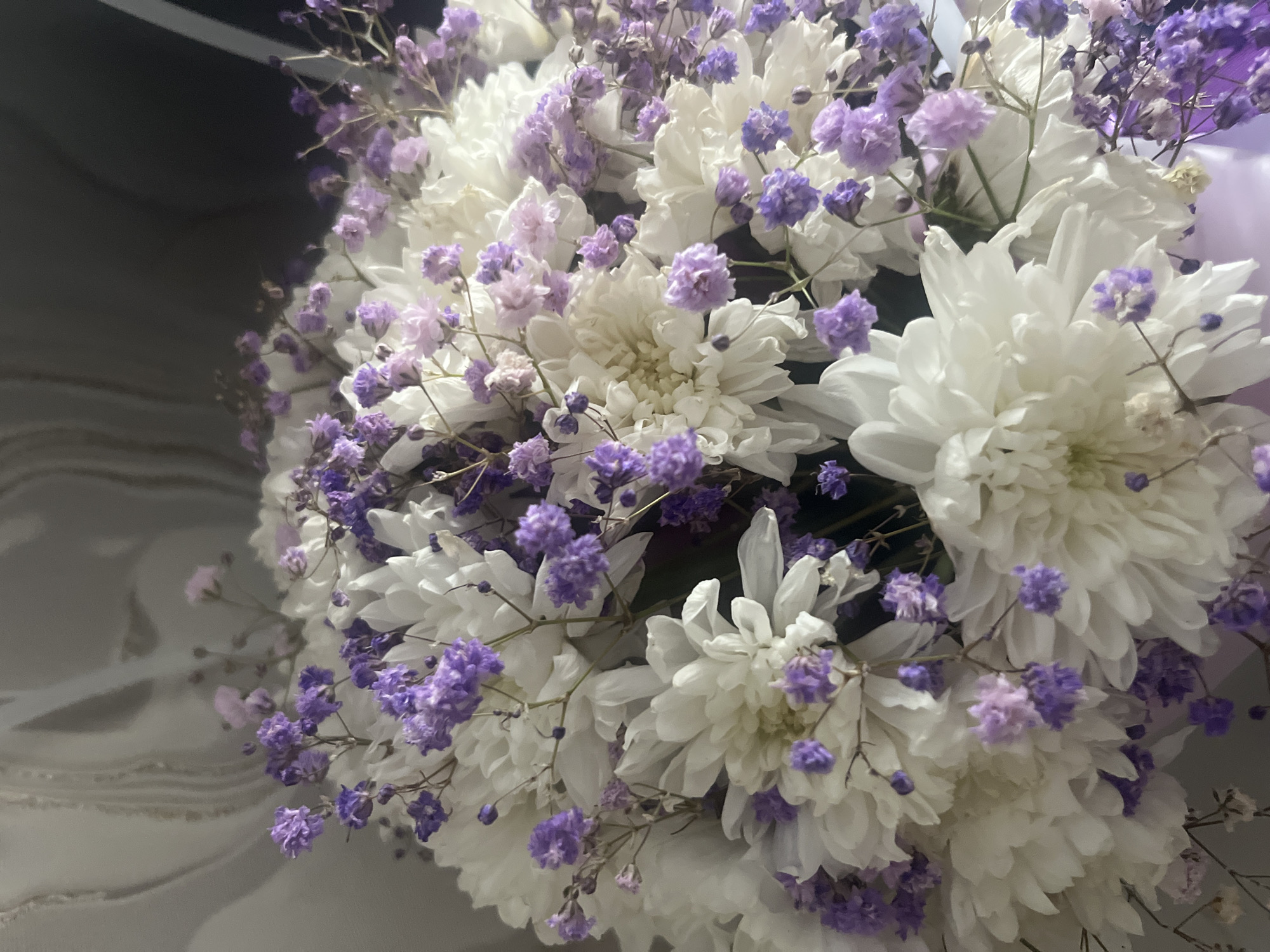 A close-up, overhead shot captures a bouquet of white chrysanthemums and small purple flowers. The background is blurred, showing a textured, light-colored wall with faint, wavy patterns, possibly wallpaper. The lighting is soft, casting subtle shadows. There are no people or animals visible in the image. The scene appears to be indoors, likely a domestic setting, but there are no specific elements to pinpoint its location within the city of Tyumen, Russia, or any other city.