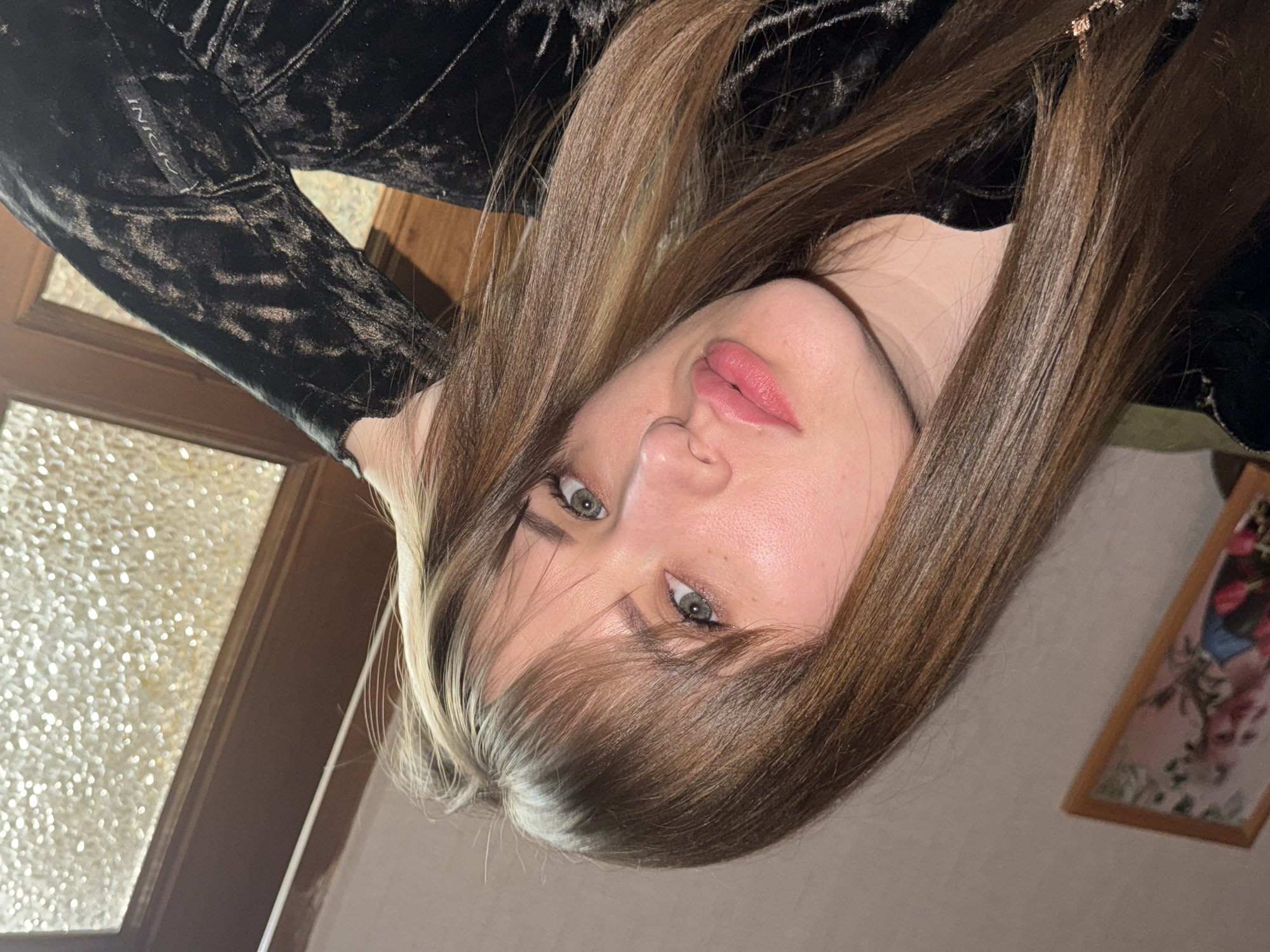 This image, taken in Chaikovsky, Russia, features a young woman, likely in her late teens or early twenties, in an indoor setting. The photograph is oriented sideways, with her head positioned towards the left of the frame. She has long, straight brown hair with lighter, possibly blonde or streaked, sections framing her face, particularly visible near her forehead. Her prominent eyes are green or blue-green, accentuated by light makeup, and her lips are softly parted with pink lipstick. Her complexion is fair, and her expression is neutral to slightly pensive.
She is wearing a dark, possibly black, velvet garment, which appears to be a top or dress with long sleeves. A visible tag on her sleeve clearly displays the text "INICIO".
The background reveals typical indoor elements. To her left, there's a wooden frame enclosing a textured, translucent glass panel, suggesting a door or window. To her right, a plain, light-colored wall is visible, with a small, framed picture depicting blurry colorful shapes, possibly flowers, further in the background. The lighting is soft, consistent with an indoor environment, and does not indicate a specific time of day. No particular activity or event is taking place; the image is a portrait focused on the woman.