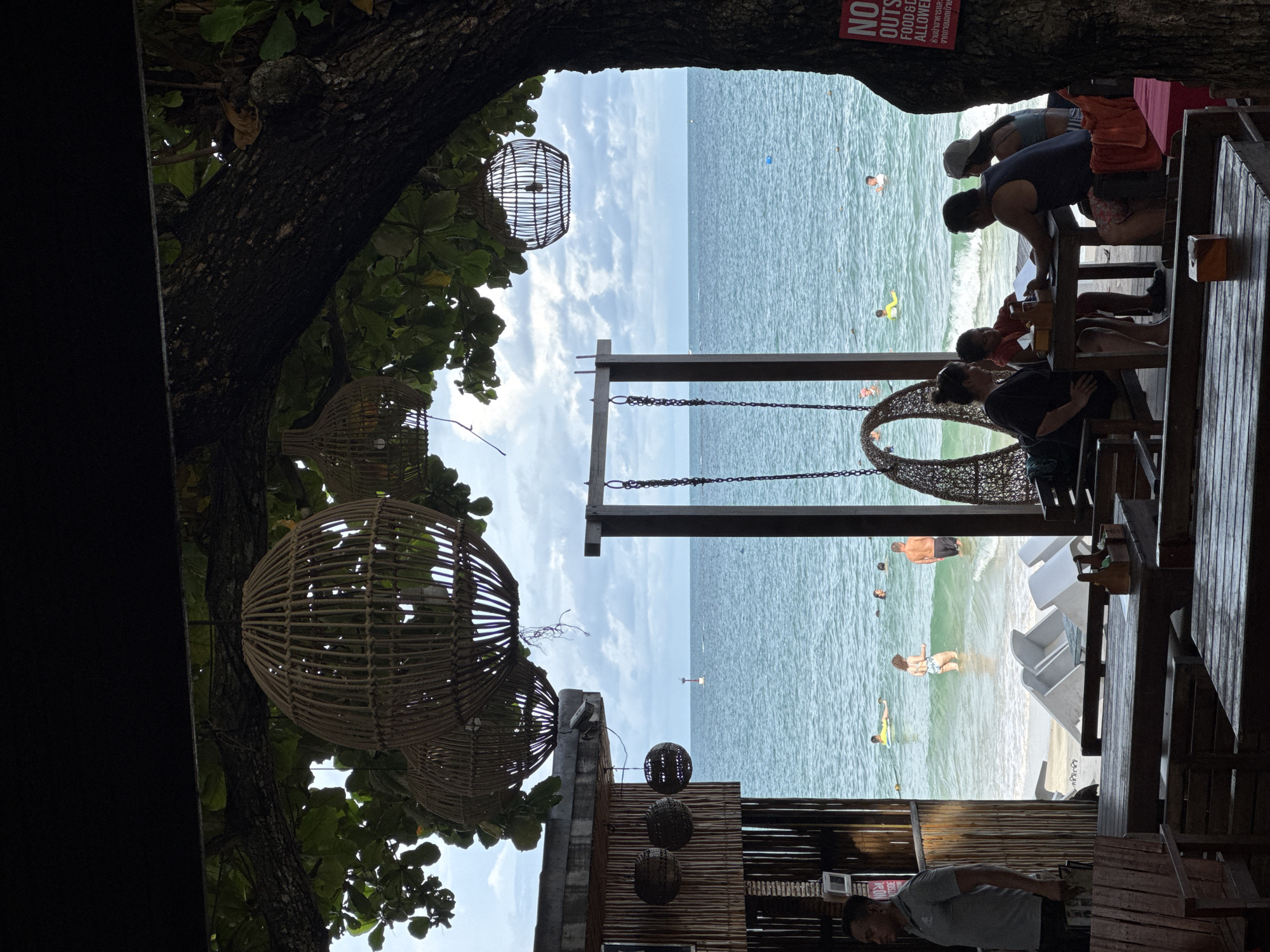 This is a medium shot of an outdoor seating area at a beachfront establishment in Ao Phai, Thailand. The foreground features wooden tables and chairs, some with cushions, set on a wooden deck. A man in a grey polo shirt stands to the left, looking towards the sea. Behind the seating area is a sandy beach with several people swimming and wading in the clear blue ocean. In the mid-ground, a wooden swing set with woven seats hangs over the sand. Overhead, decorative rattan pendant lights are suspended from tree branches. A large tree trunk dominates the right side of the frame, its branches providing shade. The background shows the ocean meeting a cloudy sky, with a distant red and white buoy visible on the water. A sign on the tree trunk reads "NO OUTSIDE FOOD & DRINK ALLOWED".