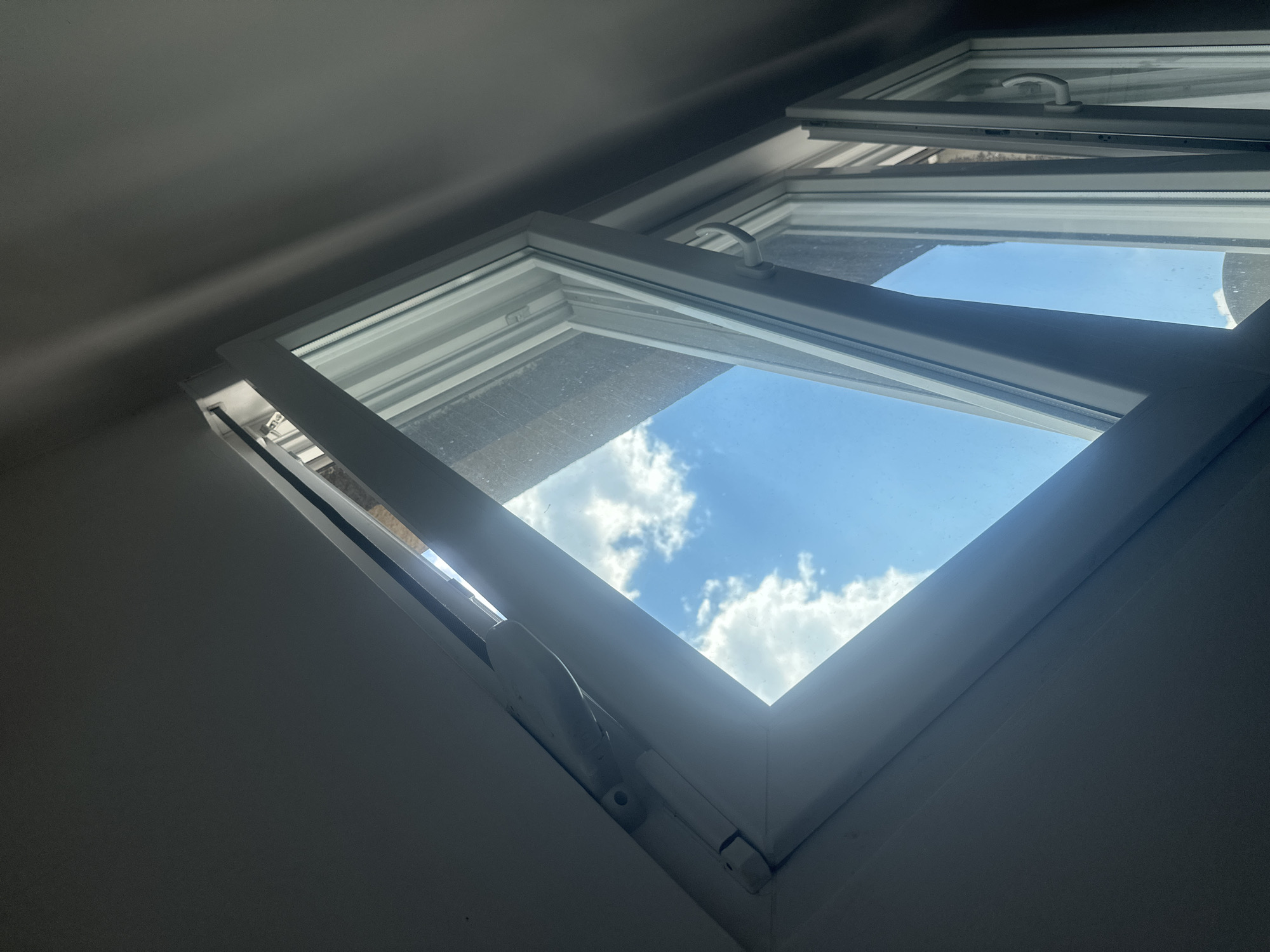 The image shows an interior view of a white-framed casement window that is partially open. The window panes reflect a bright blue sky with fluffy white clouds, suggesting it is daytime and the weather is clear. The interior walls are a neutral, dark grey color, with one section of the wall prominently lit by sunlight streaming through the open window. This creates a strong contrast between the illuminated and shadowed areas, adding depth and a dramatic feel to the scene. The window mechanism, including a white handle and hinges, is visible. The scene appears to be an indoor space, possibly a room with a skylight or an attic window, given the angle of the light and the close-up perspective. There are no people or animals visible in the image. The overall impression is one of peaceful observation of the outdoors from a dimly lit interior. The location context of Zvezdara Urban Municipality, Serbia, provides a geographical setting, but no specific visual cues within the image directly confirm this location. There is no visible text in the image.