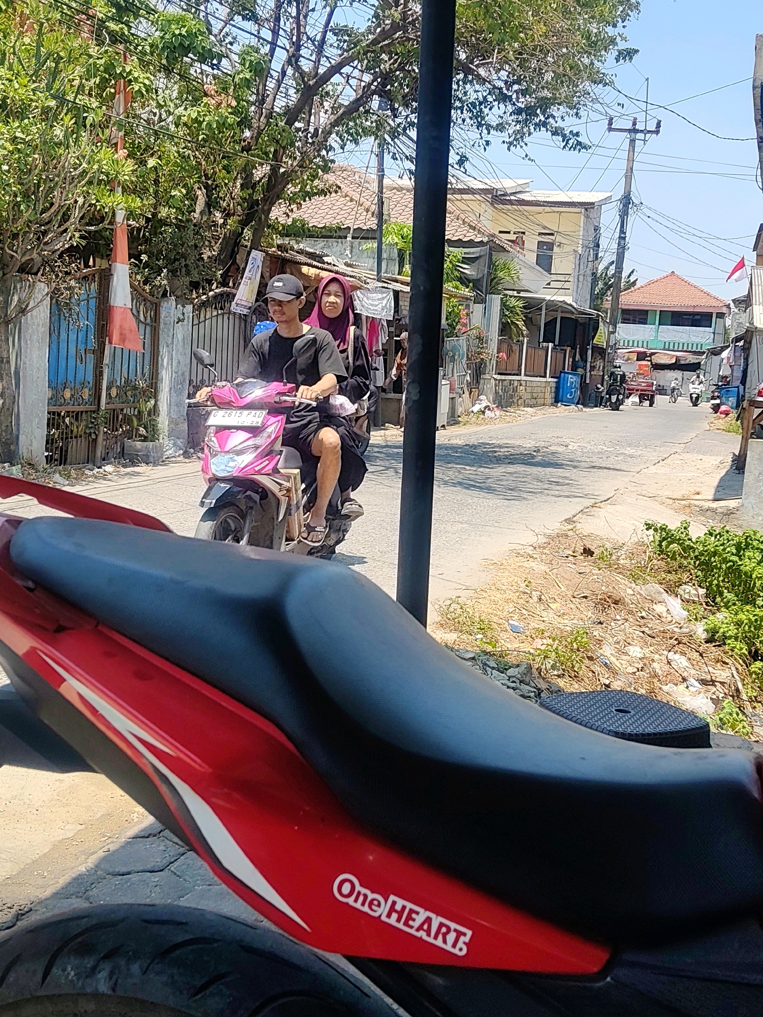 The image depicts a street scene in Mekarmukti, Indonesia, on a sunny day. In the foreground, a red motorcycle is partially visible. Further down the paved street, a pink scooter is carrying two people. The rider, a young man wearing a black t-shirt and a black cap, is operating the scooter. Behind him, a woman wearing a purple hijab and a dark dress is seated. The street is lined with houses and fences, with lush green trees overhead. Electrical wires are strung across utility poles, and there is some scattered debris along the roadside. The overall impression is one of a typical, somewhat informal urban environment.