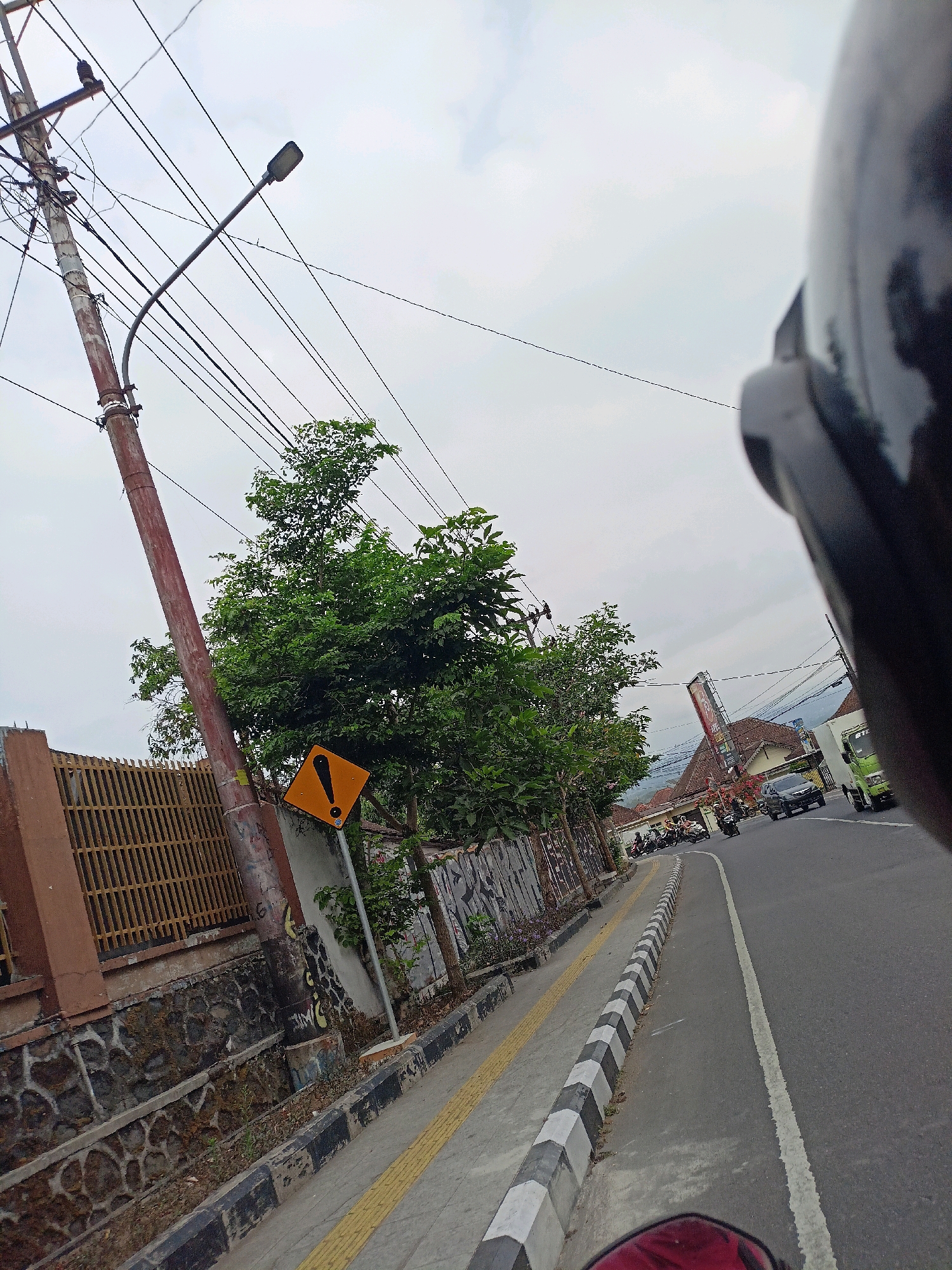 This image captures a street scene in Magelang, Indonesia. The perspective suggests the photo was taken from the viewpoint of someone riding a motorcycle, with the front of a dark helmet visible in the lower right corner.
The scene depicts a road with a distinct curb that is painted with alternating black and white stripes. To the left of the road, there is a sidewalk with a tactile paving yellow line. Beyond the sidewalk, a stone wall and a metal fence enclose a property lined with trees. A tall utility pole with numerous wires and a street lamp is prominent on the left side of the frame. A diamond-shaped yellow warning sign with a black exclamation mark is attached to this pole.
Further down the road, several motorcycles and at least one car are visible, suggesting moderate traffic. Buildings with tiled roofs line the right side of the road in the distance, along with a tall sign that has some text on it, though it is not clearly legible. The sky is overcast, indicating a cloudy or possibly rainy day. The overall atmosphere is that of a typical day on a busy street in a Southeast Asian town. The time of day appears to be daytime, given the visibility. No specific emotions are conveyed, and the focus is on the environmental details of the location.