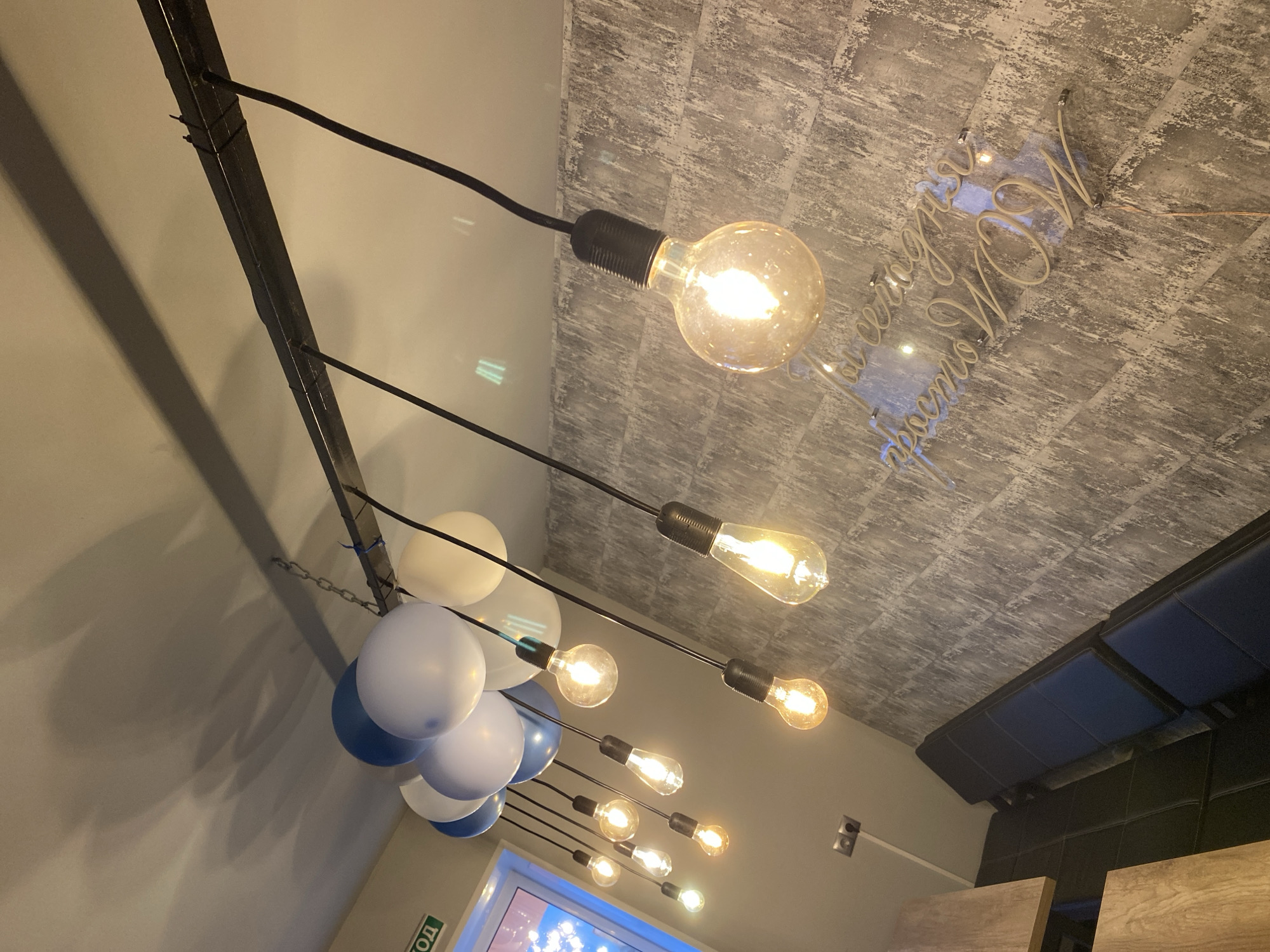The image shows an interior space with industrial-style lighting. A metal beam runs along the top left of the image, from which pendant lights hang with Edison-style bulbs. To the left, a cluster of blue and white balloons is present. The ceiling has a textured gray pattern, and a clear sign with the word "WOW" in Cyrillic is attached to the ceiling. The bottom of the image shows a window with a bright light coming through. To the right, a dark blue padded bench is visible, and wooden panels are partially visible. Based on the context clues (the Cyrillic sign), this image was captured in Chaikovsky, Russia. The scene appears to be a restaurant or cafe interior.