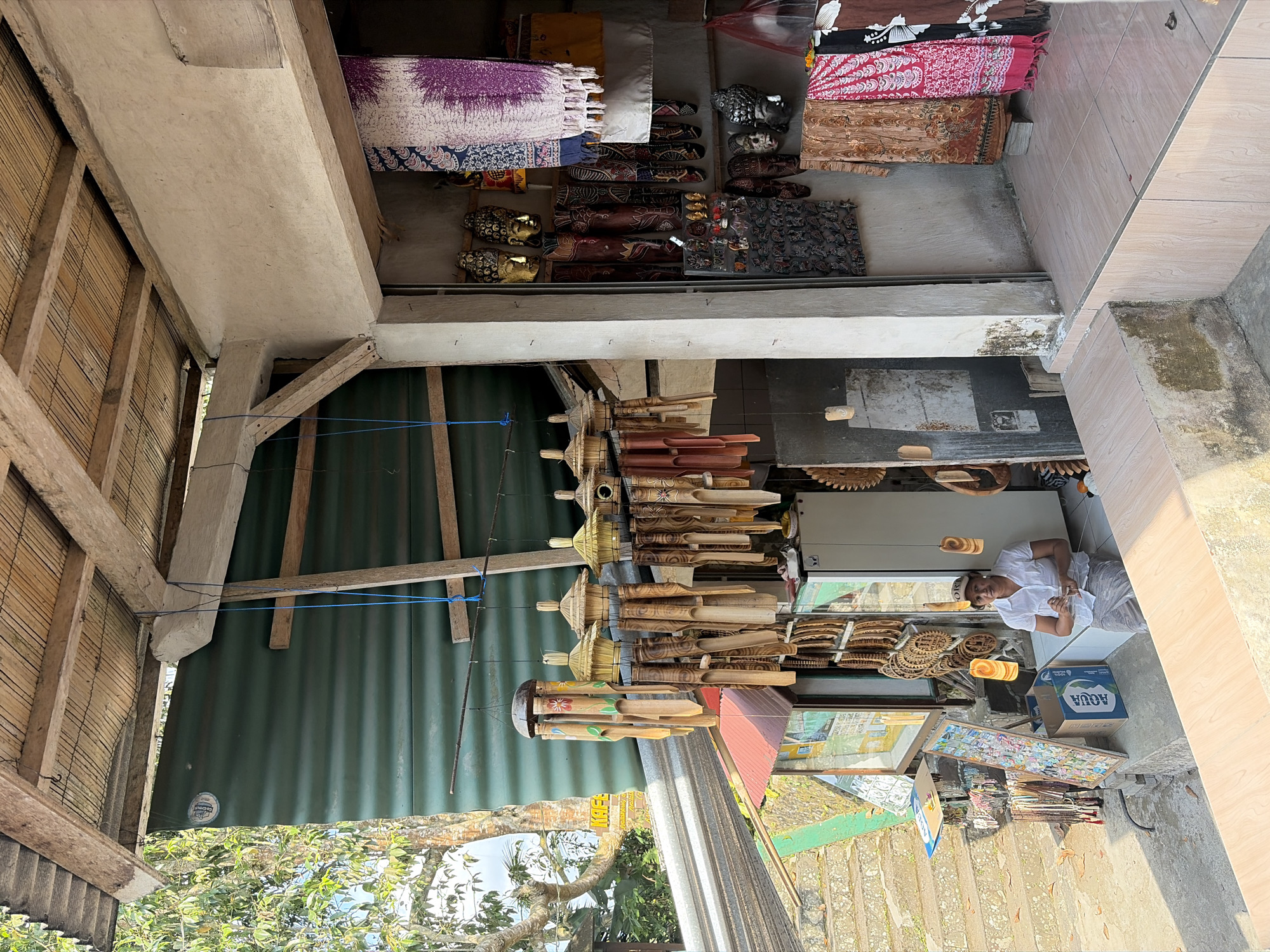 A woman sits in front of a shop in Tampaksiring, Indonesia. She is wearing a white shirt and a patterned skirt, with a beige baseball cap. She is holding a small white object in her right hand. The shop is filled with various items for sale, including wind chimes made of bamboo, carved wooden masks, and textiles. The shop is open-air, with a thatched roof supported by wooden beams. In the background, there are steps leading up to the shop, and lush green trees. The overall scene suggests a vibrant local marketplace.
