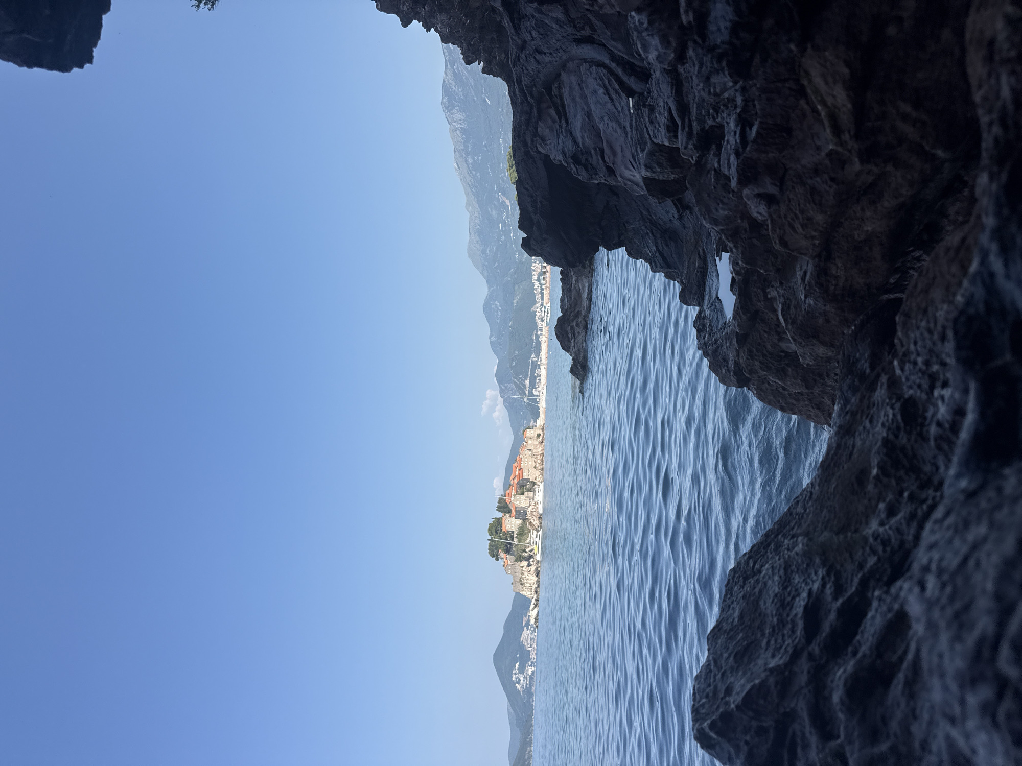 This image depicts a scenic coastal view in Sveti Stefan, Montenegro. In the foreground, dark, rugged rocks dominate the lower portion of the frame, with a glimpse of the blue water peeking through them. The middle ground features the calm, blue Adriatic Sea, with gentle ripples reflecting the clear sky.
Dominating the mid-distance is the iconic Sveti Stefan island, a small islet connected to the mainland by a narrow causeway. The island is covered in traditional stone buildings with terracotta roofs, nestled amongst lush green trees. Several sailboats are visible anchored near the island.
In the background, a range of rolling green hills and mountains form the backdrop, with a distant town or village visible along the coastline. The sky above is a vibrant, cloudless blue, suggesting a sunny day. The overall scene is peaceful and picturesque, highlighting the natural beauty of the Montenegrin coast.