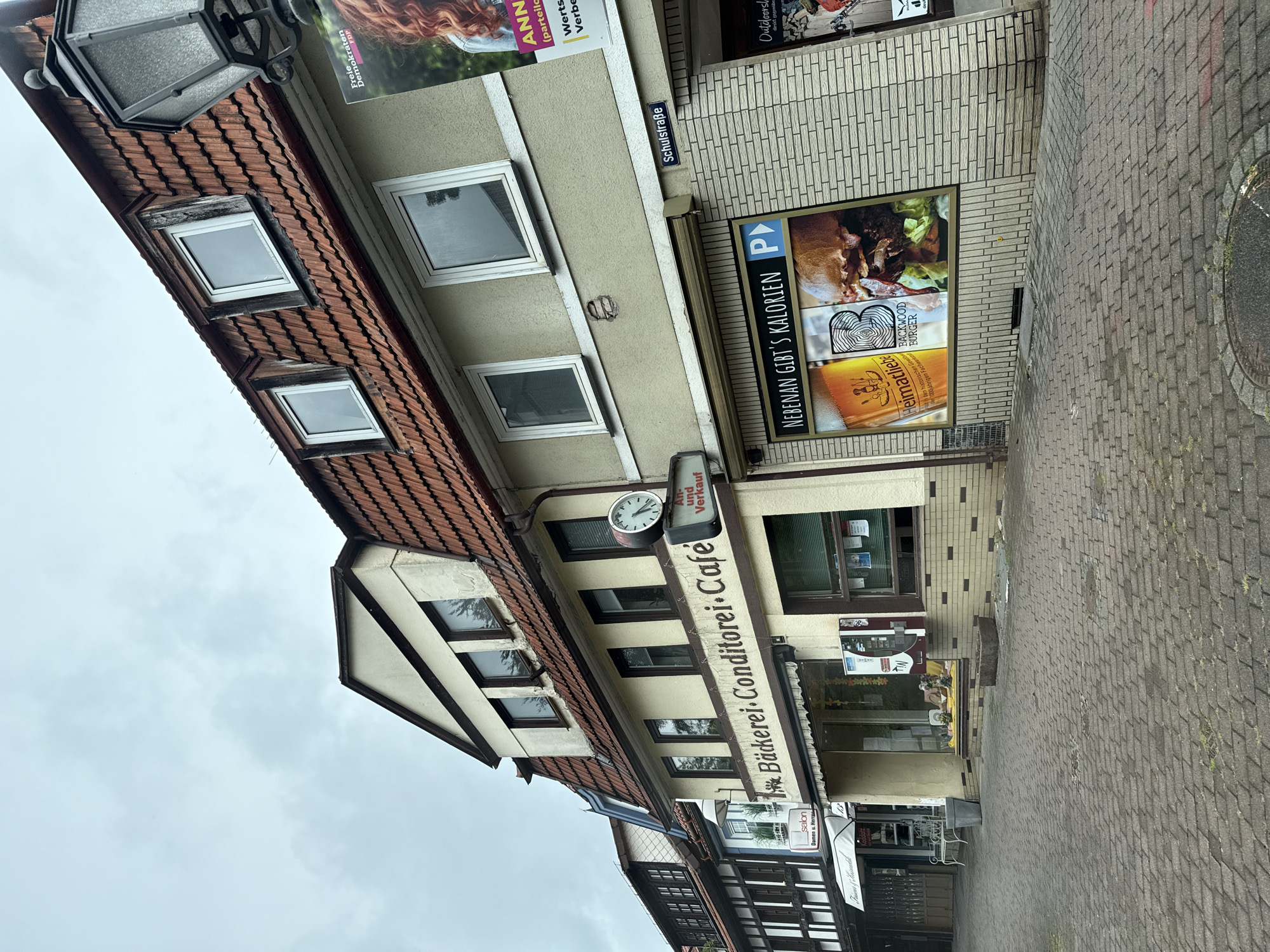 The image depicts a street scene in the town of Bad Sachsa, Germany, under an overcast sky. The foreground is dominated by a paved walkway made of interlocking bricks. On the right side of the street, a multi-story building features a light-colored facade with dark-trimmed windows and a red tile roof. A prominent sign above a storefront reads "Bäckerei-Conditorei-Café" in brown lettering. Adjacent to this is a clock mounted on the building. Further to the right, a poster advertises "NEBENAN GIBT'S KALORIEN" with an image of a burger and beer, and a street sign indicates "Schulstraße". A black lamppost is visible on the far right, partially obscuring a poster for "Freie Demokraten".
To the left of the "Bäckerei-Conditorei-Café" building, another building is visible with a darker, half-timbered style facade and smaller windows. Several shops are present, including one with a sign that reads "salon Damen u. Herren". A white awning extends over one of the shop entrances, and a metal bench sits outside. Various potted plants and decorative elements are visible in shop windows. A silver public telephone booth is mounted on the right building. The overall impression is of a quiet, European town street with commercial establishments.