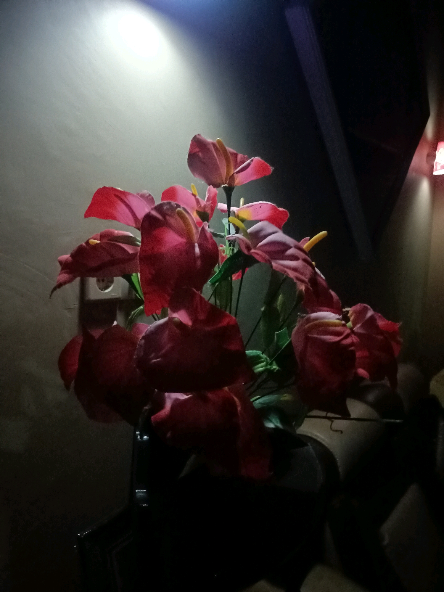 The image shows a bouquet of red and pink anthurium flowers, possibly artificial, in a dark setting. A bright light illuminates the wall behind the flowers, creating a dramatic effect. The flowers are arranged in a dark pot, which is largely obscured by shadow. The background appears to be an indoor environment, with a wall and a ceiling visible. The overall mood is somber and atmospheric. There are no people or specific landmarks visible that would indicate the location in Trenggalek, Indonesia.