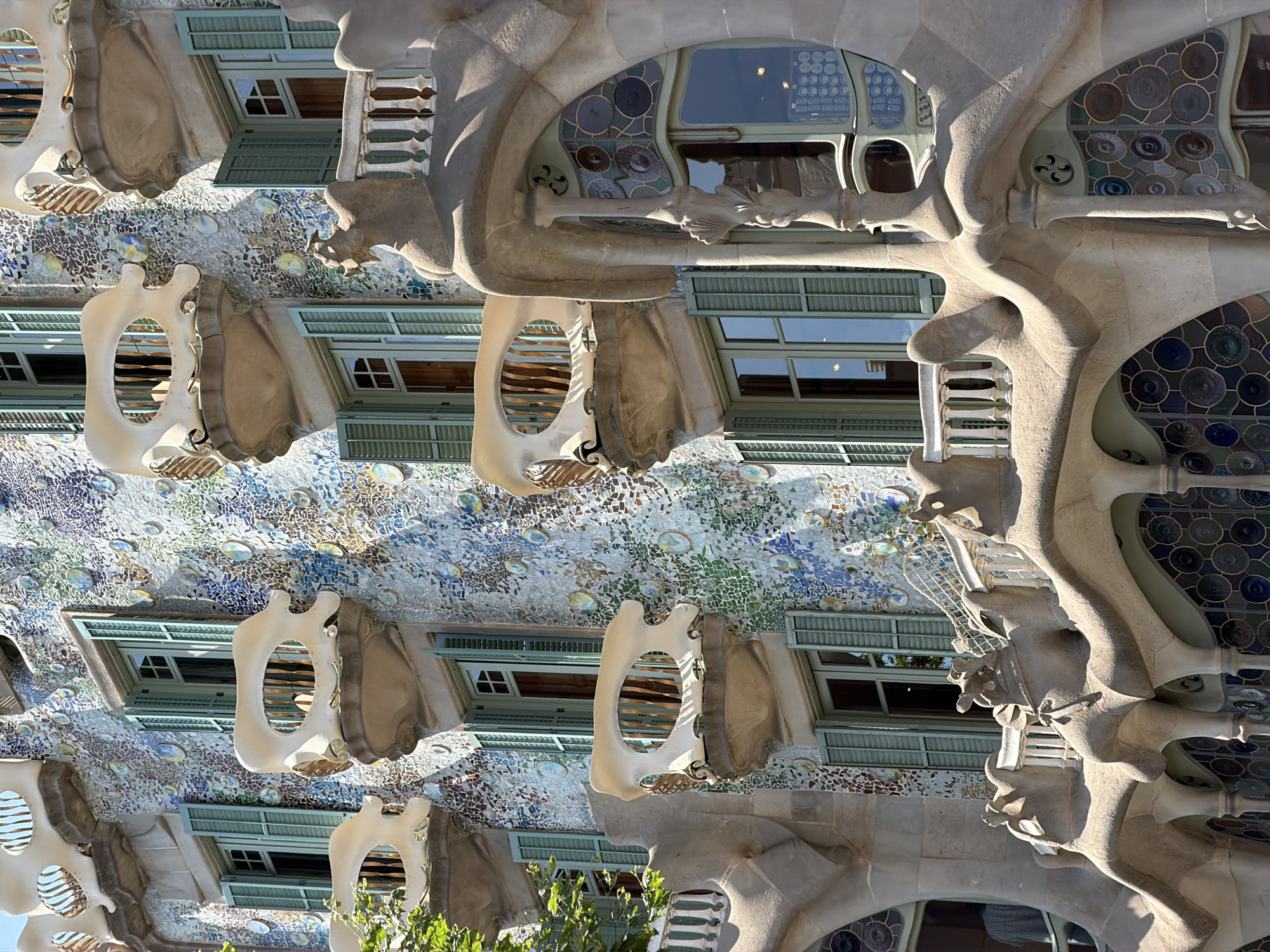 The image depicts a close-up, upward view of the facade of Casa Batlló, an iconic building located on Passeig de Gràcia in Barcelona, Spain. The building's exterior is richly decorated with colorful mosaic tiles in shades of blue, green, and white, interspersed with circular, iridescent ceramic discs.
The architectural details are a prominent feature. Several balconies, designed to resemble bones or masks, jut out from the facade. These balconies have white, sculpted ceramic fronts with oval openings that reveal wooden slatted shutters. Below these balconies, windows with light green shutters are visible.
The lower section of the building shows more organic, flowing architectural elements in a light brown stone. These sculptural forms create arched openings and decorative features. Large windows with stained glass in circular patterns are visible within these openings. In the foreground, the ornate stonework of a railing with stylized sculptures, reminiscent of mythological creatures, is visible.
There are no people visible in the image. The lighting suggests it is daytime, with bright sunlight casting shadows and highlighting the textures of the building's facade. The overall impression is one of intricate and whimsical artistry characteristic of Antoni Gaudí's work.