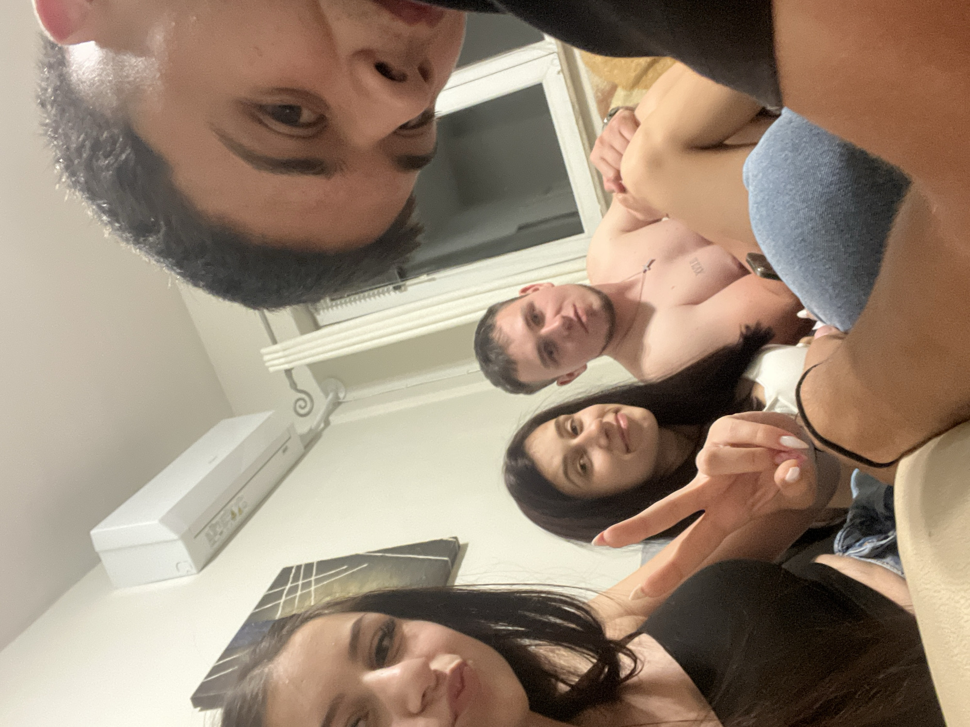 Four individuals are in an indoor environment. A man is shirtless with tattoos. A woman holds up a "peace" sign. An air conditioner is visible on the wall. A painting is also on the wall. The location is Plovdiv, Bulgaria.