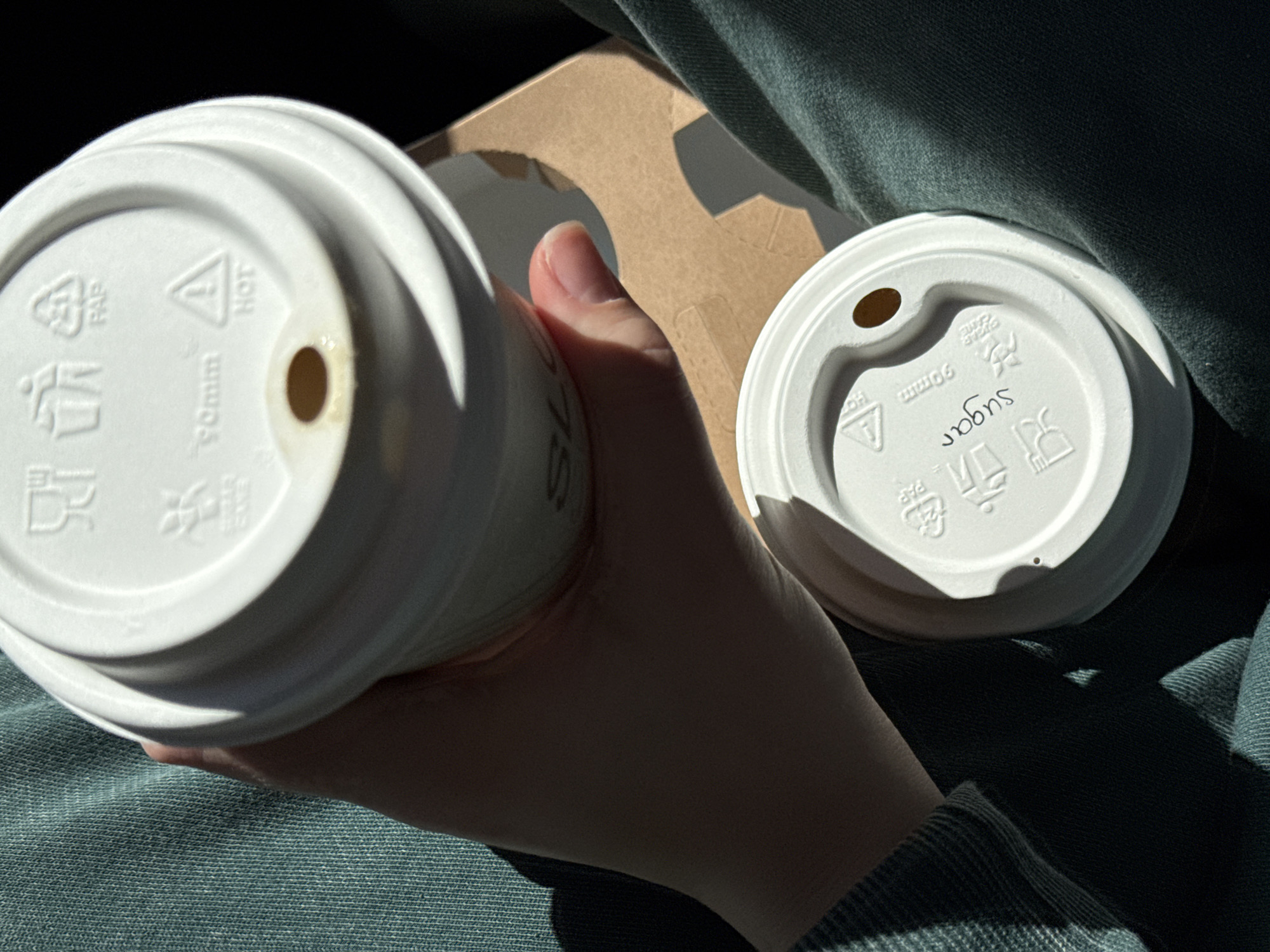 A close-up shot shows a person holding two white paper coffee cups with lids in their hand. One lid has "sugar" written on it in black pen, oriented upside down from the viewer's perspective. Both lids have small holes. The cups are positioned over a cardboard coffee carrier and the person is wearing a green sweatshirt or jacket. The lighting is bright and the location is indeterminate.