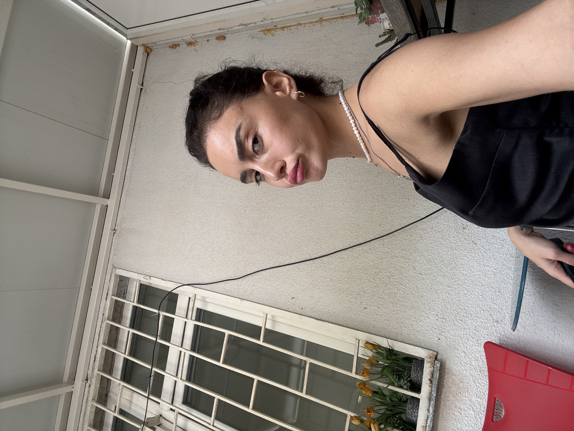 A young woman with dark hair pulled back is photographed in a selfie. She is wearing a black dress with thin straps and a pearl necklace. Her lips are pursed, and she is looking slightly to her right, away from the camera. The background appears to be an outdoor area with a textured white wall. To her left, there is a window with a metal grate and some artificial yellow flowers in planters. A red chair and a small glass-topped table are visible in the foreground. The image was taken in Tbilisi, Georgia.