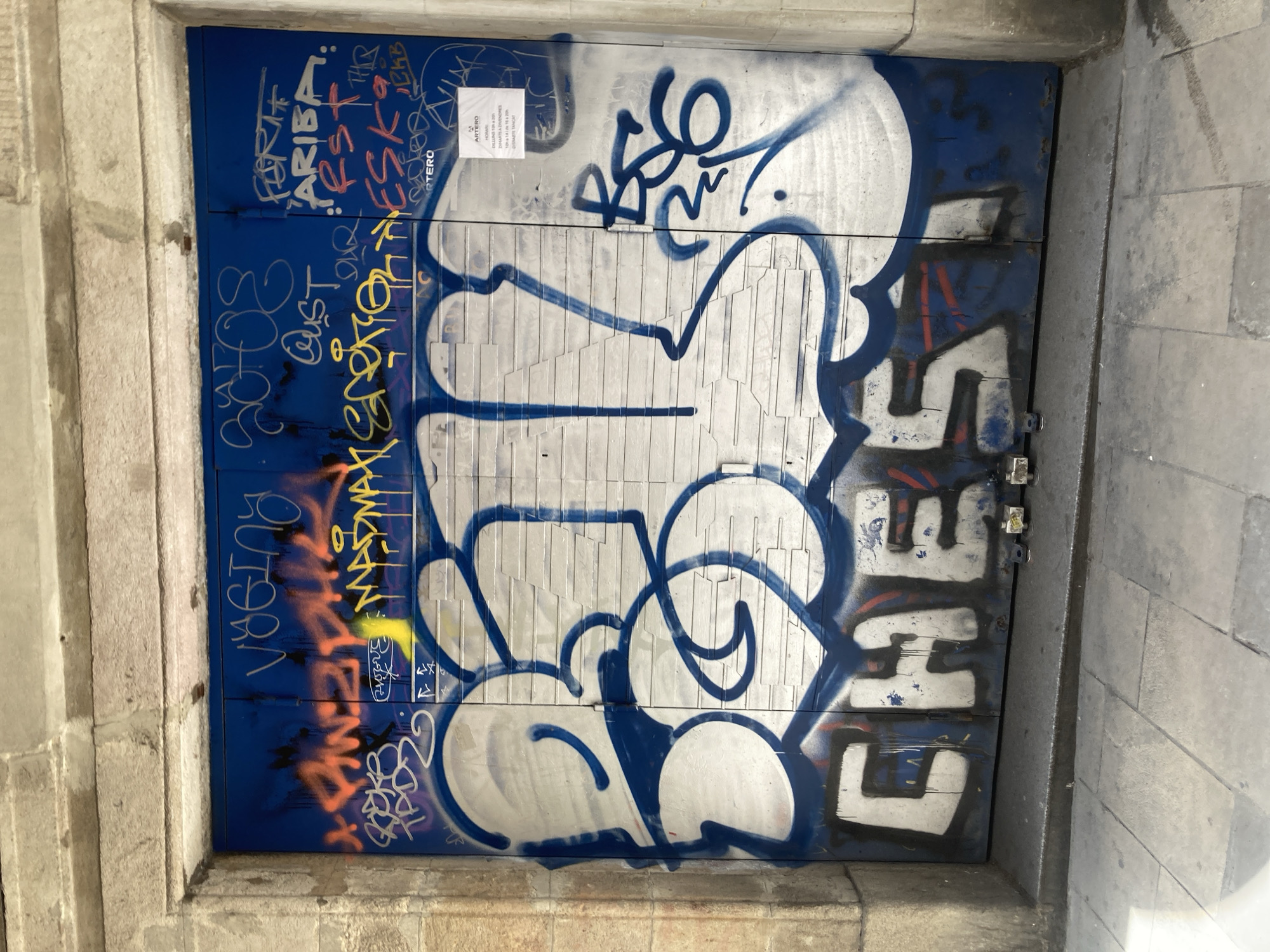 The image shows a blue metal gate in Barcelona, Spain, covered in graffiti. The gate is framed by weathered stone pillars. The graffiti is extensive and includes multiple layers and styles. Large, bubble-style letters in white with blue outlines dominate the center of the gate. Smaller tags and writing in various colors, including orange, red, yellow, and white, are visible above and around the larger lettering. Some of the visible text includes "VOGING," "MADMAX SPORT," "ARRIBA," "RST," "ESK," and "CHEST." There is also a small sign with text in Spanish that appears to be business hours. The gate is closed, and the ground in front of it is paved with gray rectangular stones.