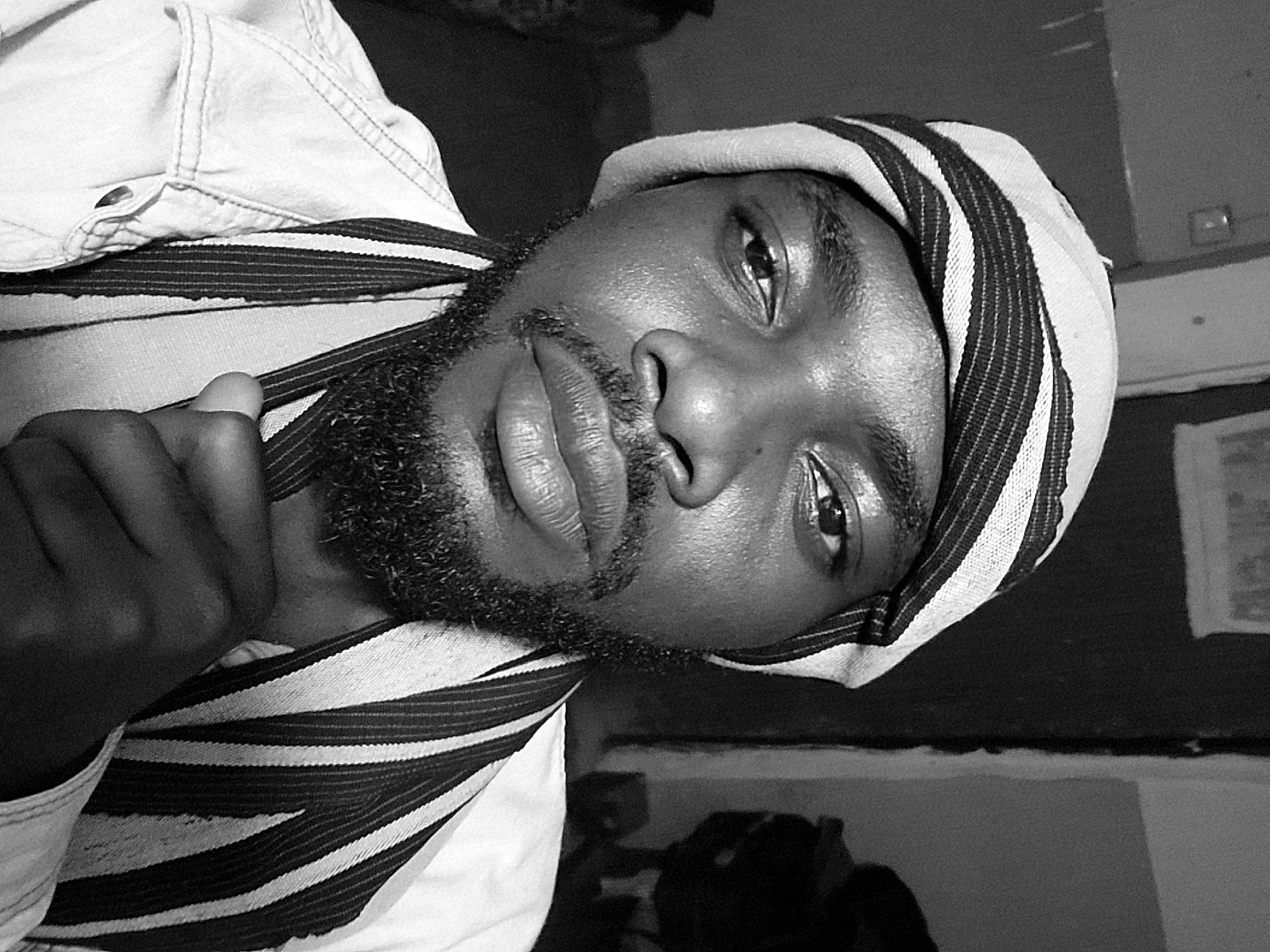A close-up, black and white portrait of a man with dark skin, a short, well-groomed beard, and a striped head covering. He's looking directly at the camera. His fingers are partially visible, near his neck. He also wears a light colored denim or cotton shirt or jacket. The background is blurry and indistinct but appears to be an interior setting. There is no evidence to support the location as Jos, Nigeria.
