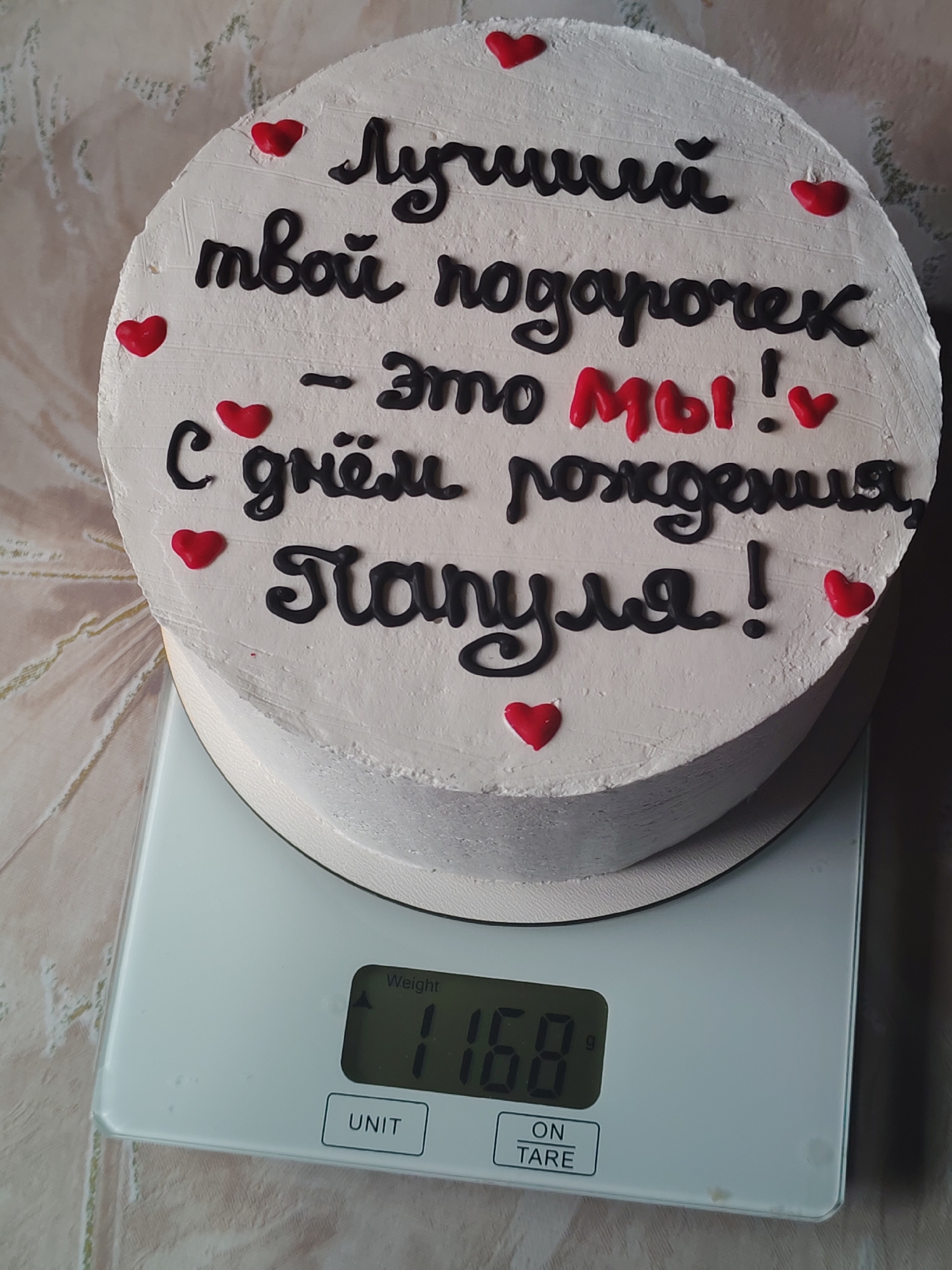A round white frosted cake sits on a digital scale that reads "1168 g". The cake is decorated with black lettering and small red hearts. The text in Cyrillic reads: "Лучший твой подарочек - это мы! С днём рождения, Папуля!" ("Best gift is us! Happy birthday, Daddy!"). The background is a light-colored, textured surface.