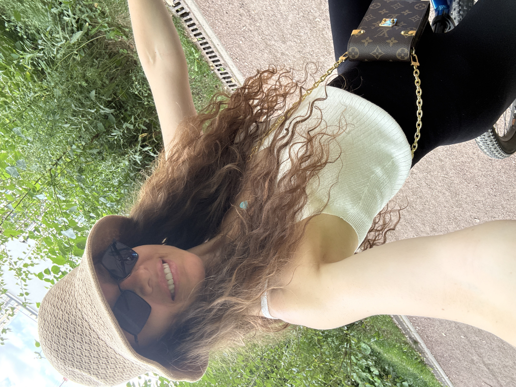 A woman wearing a light-colored knit tank top, black leggings, a straw bucket hat, and dark sunglasses is sitting on a bicycle. She is smiling at the camera, and her long, curly brown hair is visible. She has a small, brown designer purse with a gold chain strap. The background shows lush green foliage and a paved path. The image appears to be taken outdoors in a park-like setting. There is no indication that this location is in Moscow, Russia.