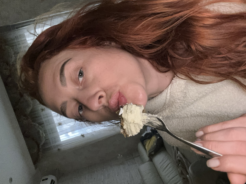 The image displays a female subject, observed from the chest up, indoors. She possesses long, reddish-brown hair and light-colored eyes, with her lips pursed into a puckering expression. She is clad in a light-colored, textured, fleece-like top. Her right hand, partially visible, holds a silver-colored fork bearing a portion of a multi-layered dessert. This dessert consists of a light-colored, creamy substance over a dark base, topped with white specks, and is positioned directly in front of her mouth. The background includes a light-colored wall, an air conditioning unit mounted in the upper left, and a partial view of a light-colored sofa with a patterned cushion. A window, draped with sheer white curtains, is situated behind the subject. The precise location within the city of Budva, Montenegro, is not discernible from the visual information present in the image.