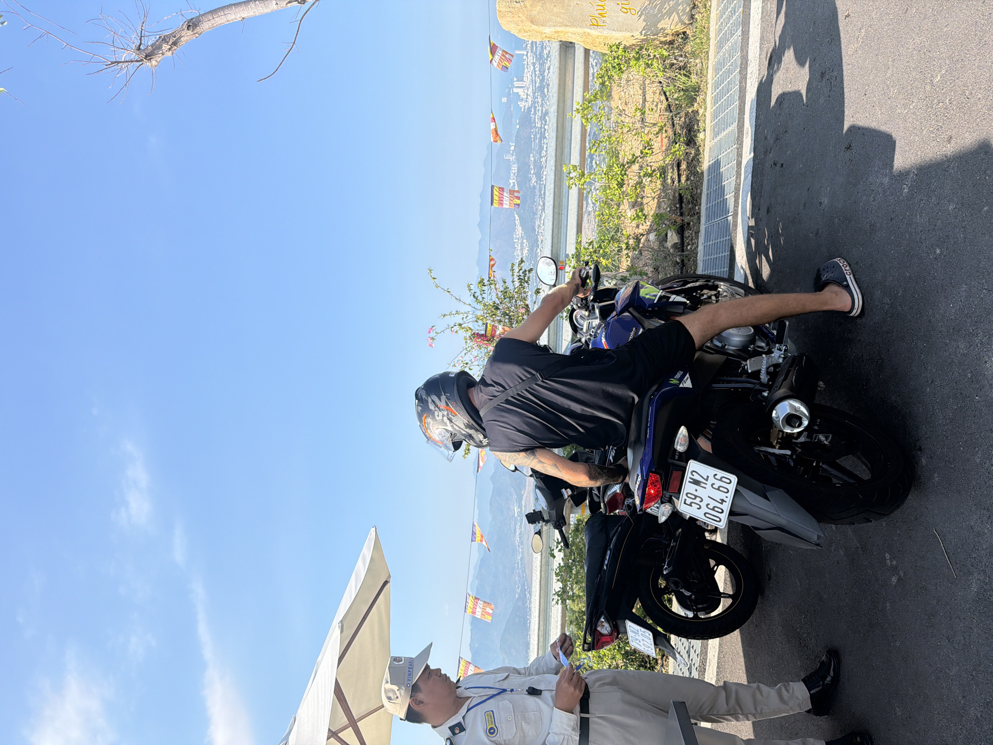 A motorcyclist wearing a helmet and black clothing is seated on a blue motorcycle with license plate number "59-M2 064.66" in Nam Nha Trang Ward, Vietnam. He's interacting with a security guard in a white uniform standing beside the motorcycle. In the background, prayer flags are strung across a scenic vista. The sky is clear and blue.