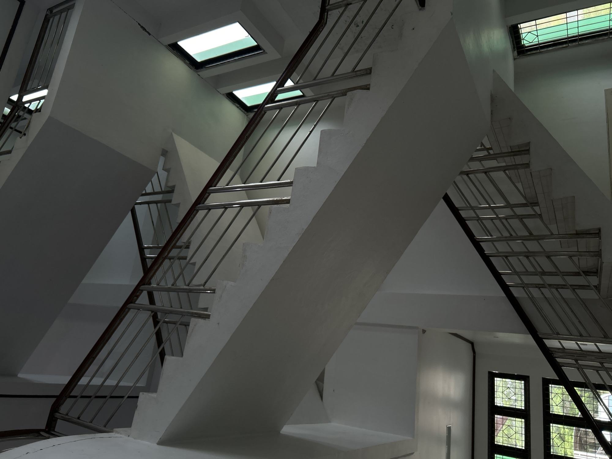 This is an interior architectural photograph capturing a complex, multi-level stairwell within a building, likely located in Reuleut Barat, Indonesia. The scene is devoid of people and activities, focusing solely on the structural elements.
The composition features several overlapping flights of stairs made of light-colored concrete or painted white. These stairs ascend and descend at various angles, creating a dynamic and somewhat disorienting geometric pattern. Robust metal railings, appearing to be stainless steel or a similar alloy, run alongside the staircases, providing safety and continuity. Some sections of the railing appear darker, possibly due to shadows or a different material.
Illumination comes from multiple sources. Rectangular light fixtures, possibly skylights or recessed ceiling lights, emit a bright, cool light from above, highlighting the clean lines of the architecture. Additionally, natural light enters through gridded windows with dark frames visible in the upper and lower right sections of the image, revealing lush green foliage outside, suggesting it is daytime. The overall palette is dominated by whites, grays, and metallic tones, with touches of bright green from the exterior. The interplay of light and shadow emphasizes the building's structural forms and creates depth. No visible text is present in the image.