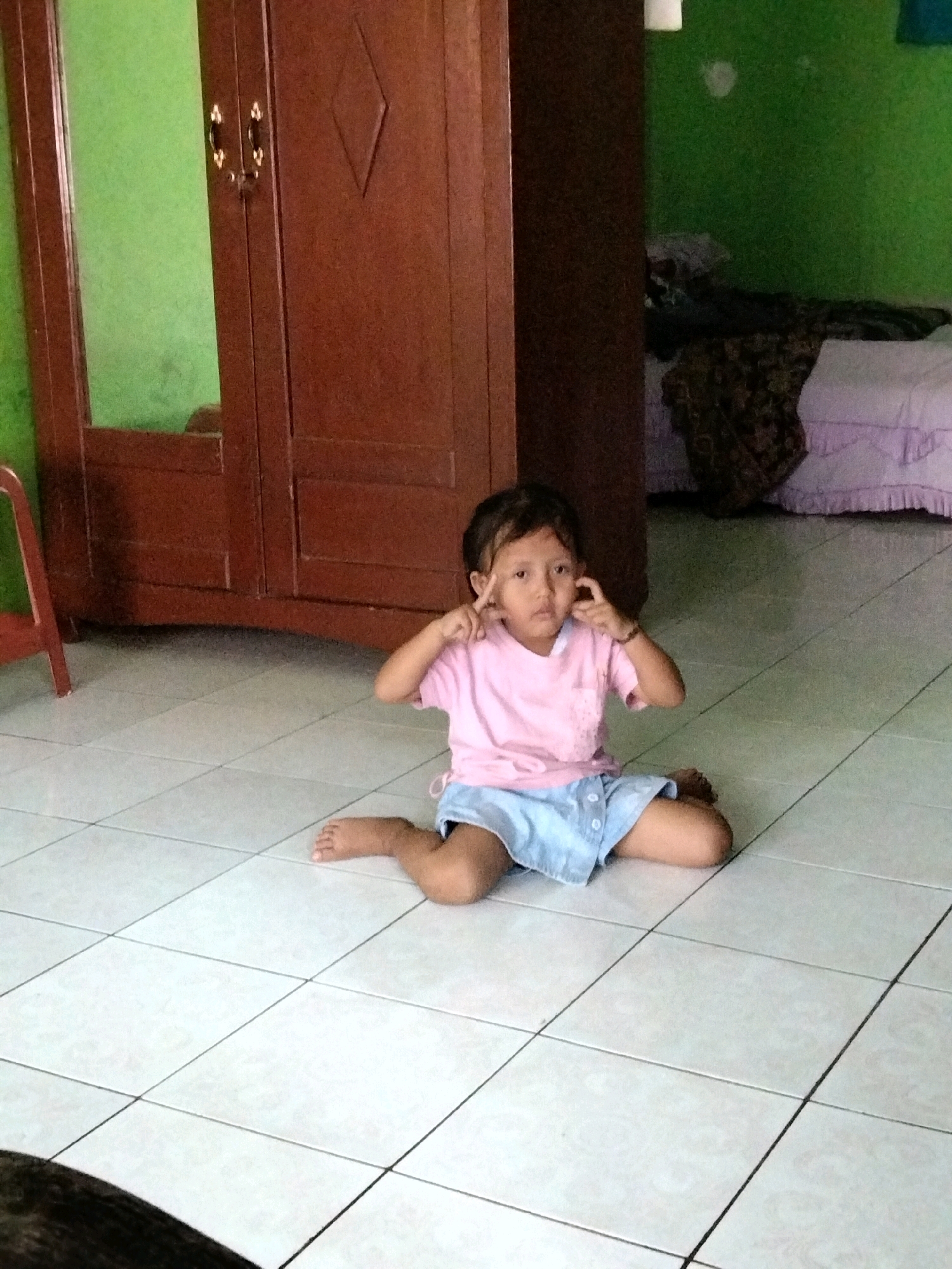 A young girl with dark hair is sitting cross-legged on a tiled floor, her knees bent and her feet pointing to the right. She is wearing a light pink t-shirt and a light blue skirt. Her hands are raised to her face, with her index fingers touching her temples, as if she's thinking or posing for a picture. She has a neutral expression.
Behind her, to the left, is a large, dark wooden wardrobe with two doors and a diamond-shaped detail on the right door. A mirror is visible on the left door, reflecting a green wall. To the right of the wardrobe, a bed is visible with white sheets and a patterned throw blanket. The wall behind the bed is also green.
The lighting suggests it is daytime. The scene appears to be an indoor setting, possibly a bedroom or living area in a home, given the wardrobe and bed. The tiled floor is white with dark grout lines. There is no visible text in the image. The overall atmosphere is casual and domestic.