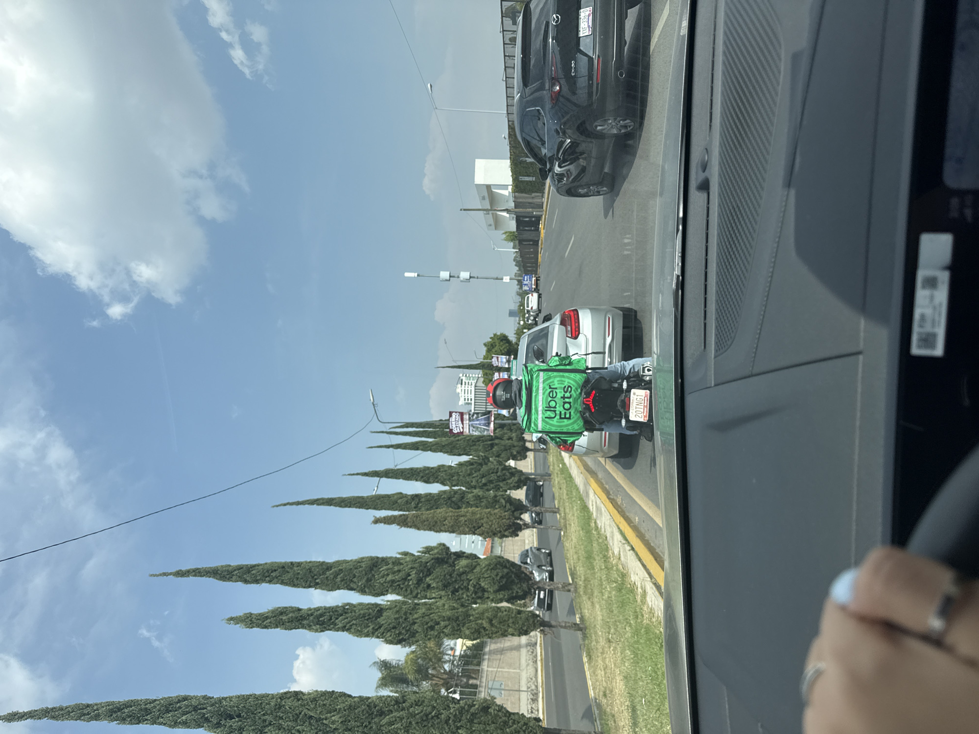 The image, taken from inside a car, shows a scene on a road in Puebla, Mexico. An Uber Eats delivery person on a motorcycle is in the adjacent lane. The motorcycle's license plate number is 20TNG1. The sky is blue with scattered clouds, and a line of tall, thin trees lines the side of the road. Other vehicles are present on the road, including a silver car and a black SUV. The dashboard of the car from which the photo was taken is visible, with a hand on the steering wheel in the lower right corner.