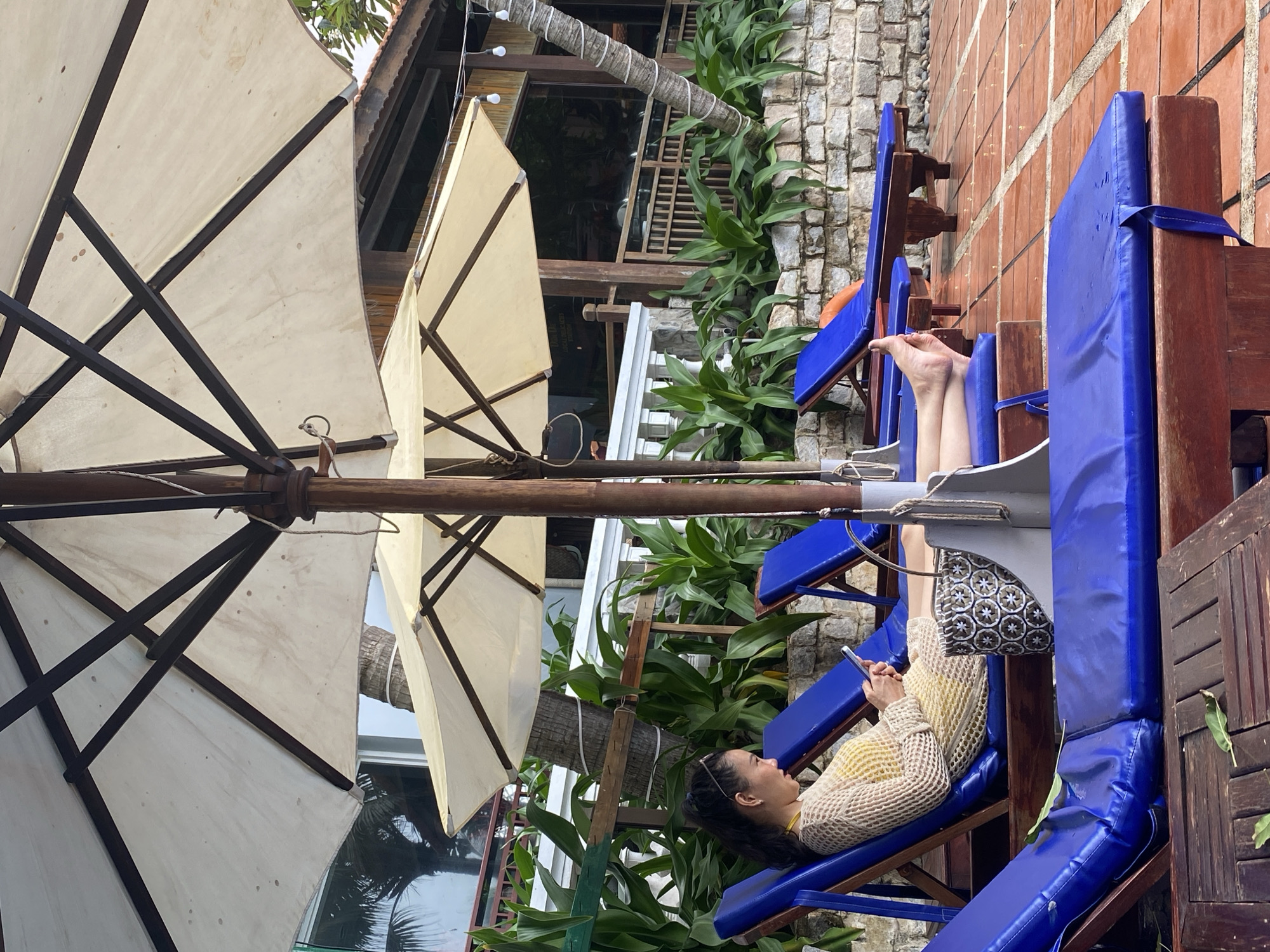 The image shows a woman lying on a blue beach chair under a large umbrella in what appears to be a resort or similar establishment in Phu Thuy Ward, Vietnam. The woman is wearing a yellow outfit with a net-like outer layer and is looking at her phone. Her legs are elevated on the chair. A patterned bag is nearby. The surrounding environment includes greenery, palm trees, a building with a wooden structure, and a stone wall. Other blue beach chairs are visible. The umbrella is large and has a tan or cream-colored canopy with dark supports.