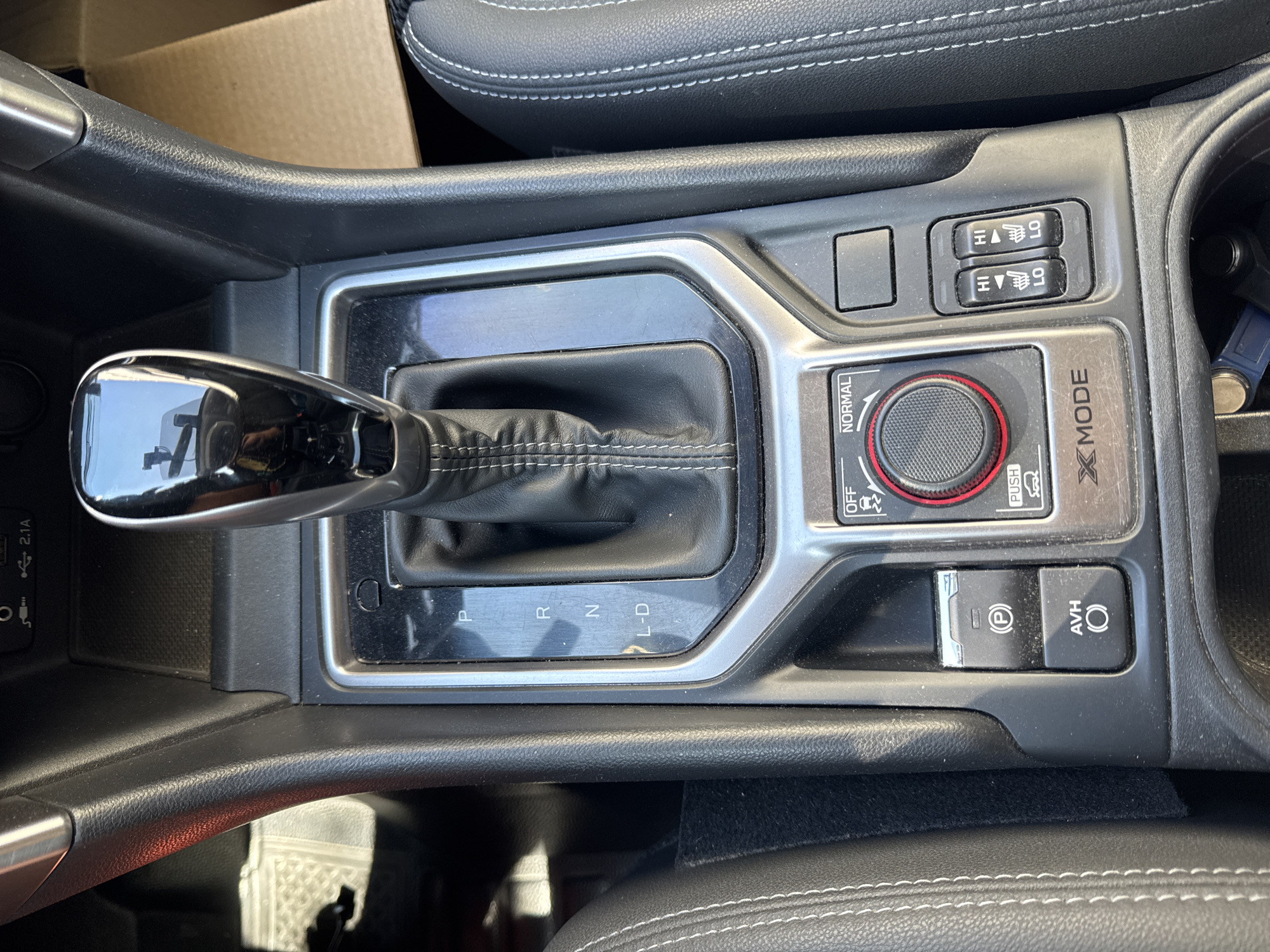 This is a top-down, close-up shot of a car's center console, featuring the gear shift, various buttons, and a dial. The gear shift is in the "P" (Park) position. To the left of the gear shift is a button with a "P" inside a circle, indicating the parking brake, and below it is a button labeled "AVH" with a circle, likely for Auto Vehicle Hold. To the right of the gear shift is a rotary dial with modes labeled "OFF", "NORMAL", and a symbol for "X-MODE". Below the dial are two sets of buttons with "HI" and "LO" markings, suggesting heated seats or climate control adjustments. The console is surrounded by black and silver trim. The overall environment appears to be inside a vehicle, with the context of the city of Barnaul, Russia, not discernible from the visual information provided in the image.