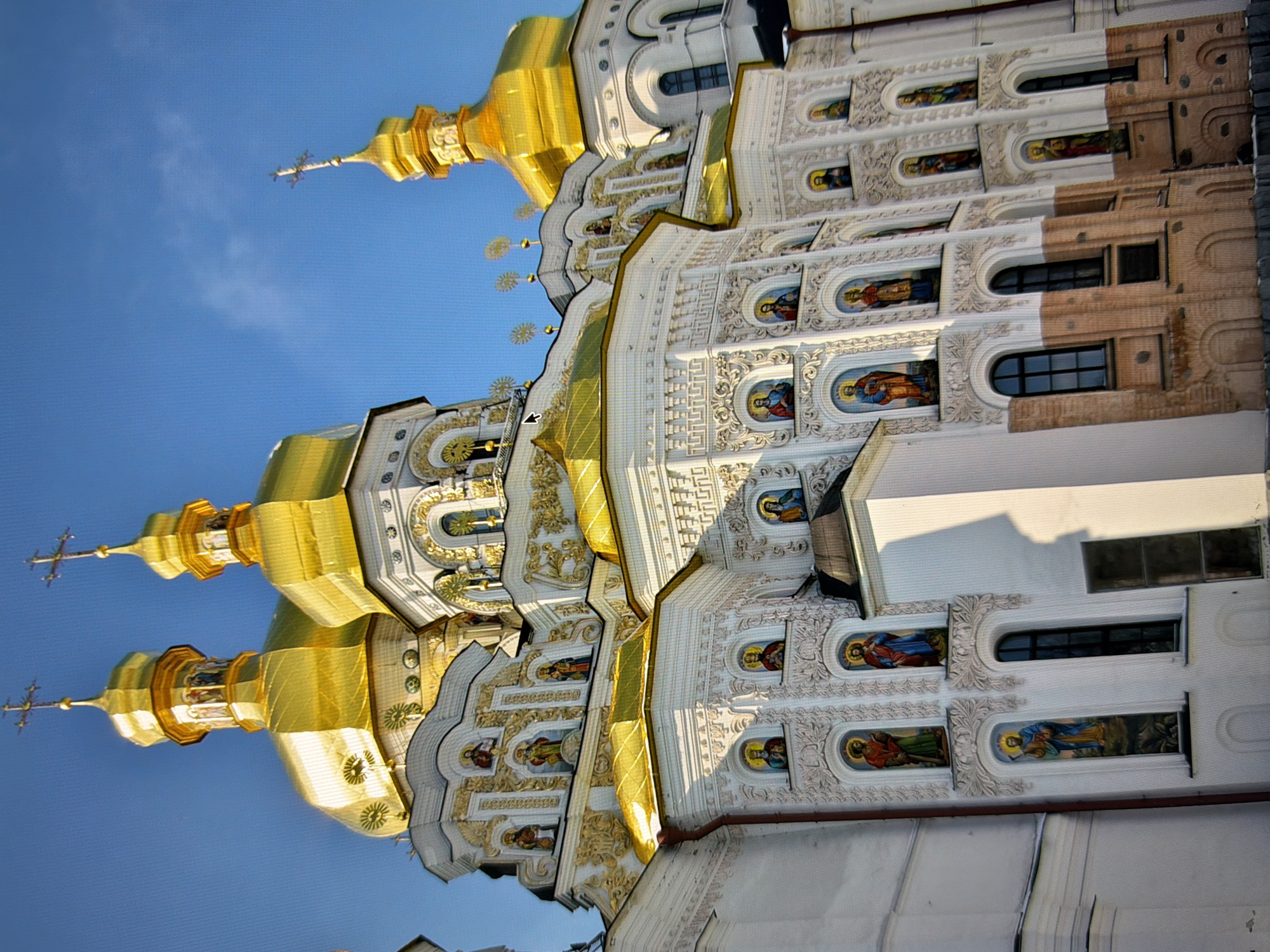 The image displays the exterior of the Kyiv Pechersk Lavra, a historic Orthodox Christian monastery located in Kyiv, Ukraine. The architecture features multiple golden domes topped with crosses, characteristic of Byzantine and Baroque styles. The white facade is adorned with intricate decorative elements and numerous religious icons depicted in arched niches. The sky is clear and blue, suggesting a sunny day. There are no people visible in the image.