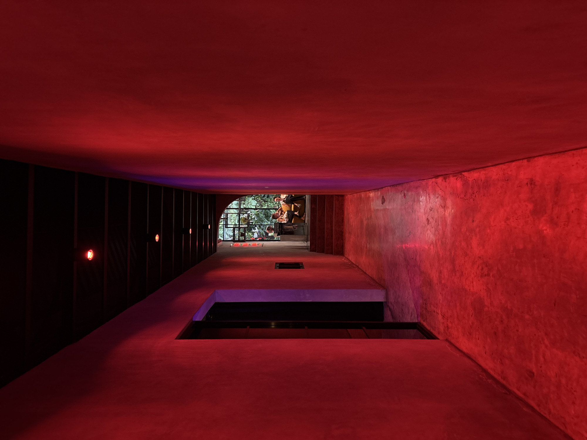This image captures a striking architectural interior, largely enveloped in a deep, vibrant red light. The scene depicts a long, seemingly enclosed corridor or a multi-level void within a building, with the perspective looking downwards and forwards. The dominant red light emanates from an unseen source, bathing the concrete-like walls, ceiling, and floor, with the right wall exhibiting a textured, somewhat rough finish. The ceiling subtly transitions to a purple hue towards the far end of the space.
On the left, a dark, panelled wall is punctuated by several circular, glowing red lights, contributing to the dramatic illumination. The foreground features a large, rectangular cutout in the red floor, revealing a darker, lower level below.
At the distant end of this red-lit passage, an arched opening frames an exterior view filled with lush green foliage, suggesting a tropical or subtropical environment. Several casually dressed individuals are visible in this brighter, outdoor area, engaged in what appears to be social interaction or relaxation—one person is seated on steps, another is reclining.
The strong, artificial lighting creates a moody and distinctive ambiance, likely intended for an evening setting in a contemporary lounge, bar, or hotel common area, fitting for its location in North Kuta, Indonesia. Despite the interior's artificial light, the visible daylight from the arched opening hints that it may be daytime outdoors. There is no visible text in the image.