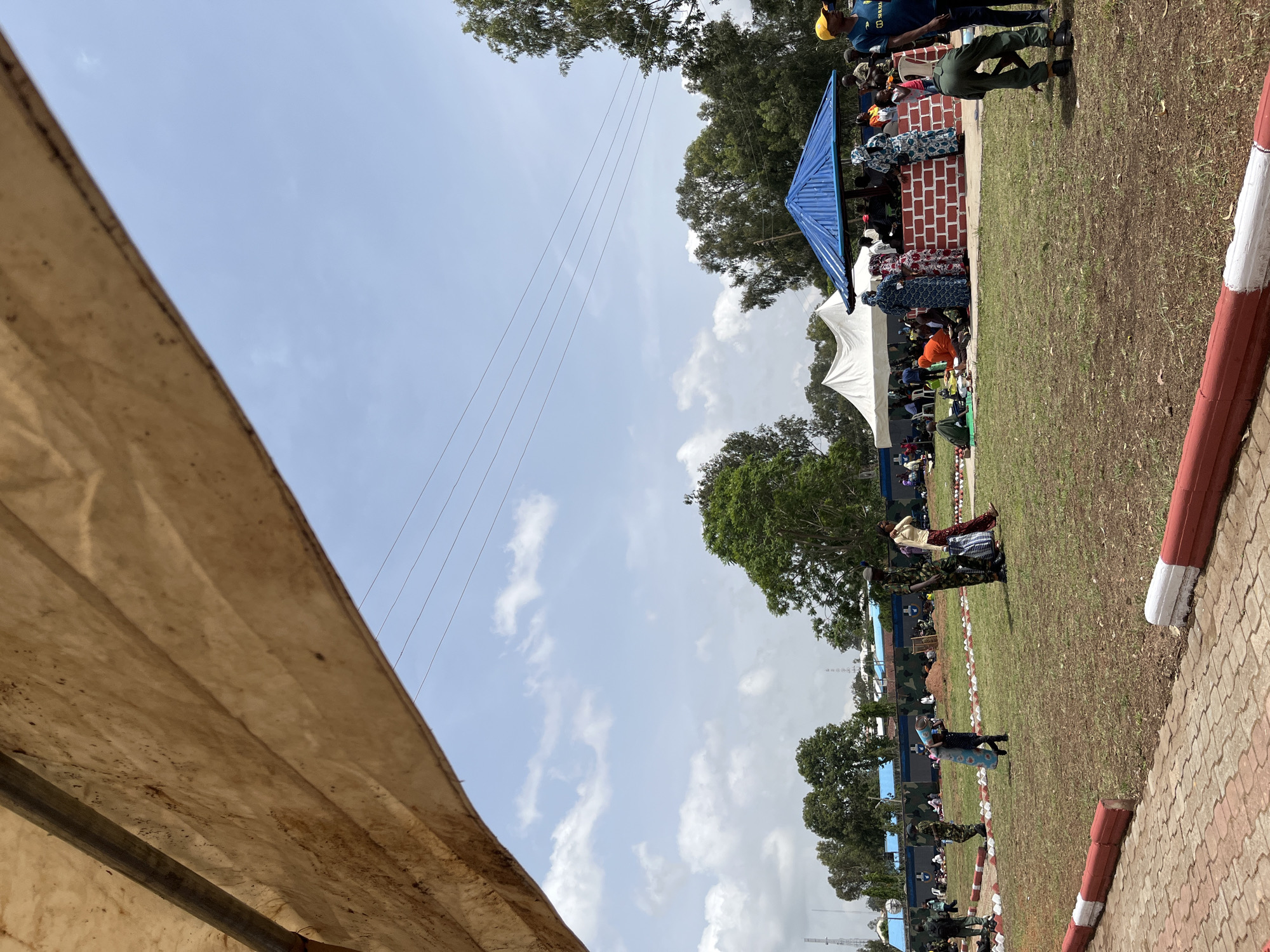 This image captures an outdoor gathering scene in the city of Bar Jirgi Summa, Nigeria. A large, tan-colored tarpaulin stretches across the top portion of the frame, partially obscuring the sky and a backdrop of trees. Below the tarpaulin, a grassy field is populated by numerous individuals. In the foreground, a paved area with red and white curbing is visible.
Several people are scattered across the field, some sitting on the ground, others standing. A group is gathered around a brick structure topped with a blue roof, under which more people are seated. White tents are set up in the middle ground, suggesting an event or gathering is taking place. Various individuals are wearing traditional Nigerian attire, while others are in military uniforms. The overall atmosphere appears to be that of a public event or communal gathering. The environment is characterized by a clear sky with scattered clouds, green grass, and numerous trees. Power lines are visible stretched across the sky in the upper portion of the image.