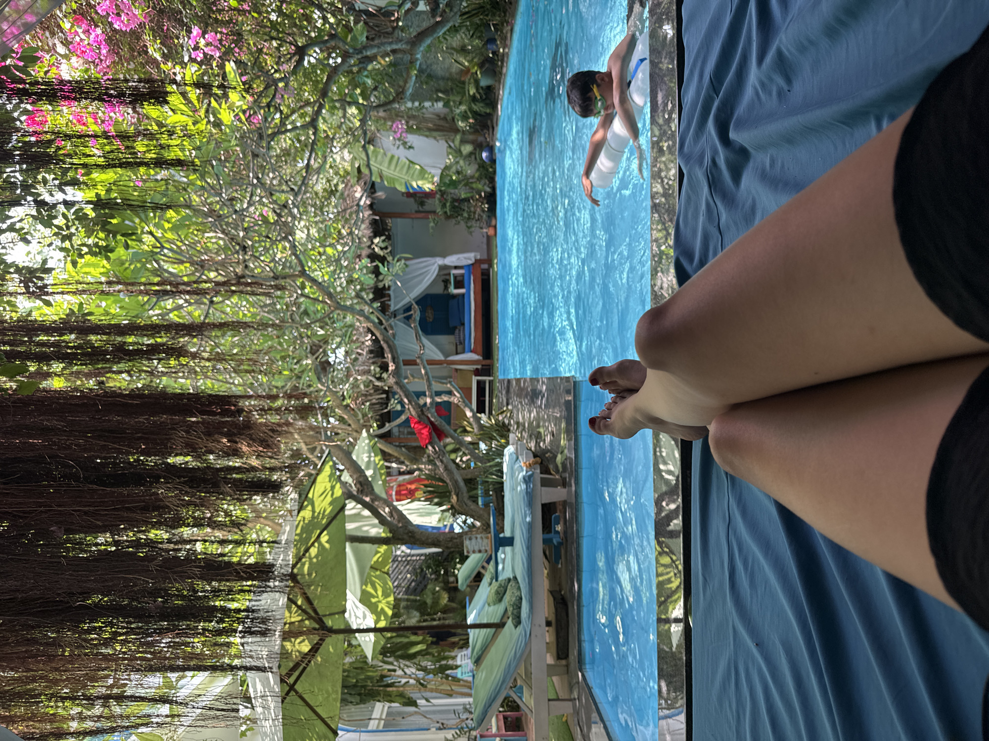 A person is reclining on a blue lounge chair next to a blue-tiled swimming pool. Another person is swimming in the pool, wearing a white top and goggles with a white inner tube flotation device. The foliage consists of lush green trees and vines hanging over the pool area, as well as pink flowers. The setting appears to be an outdoor lounge area with beach umbrellas and lounge chairs near the swimming pool. A building with a white curtain is visible in the background. While the location cannot be determined without other information, I cannot find evidence that this is in Mũi Né, Vietnam.