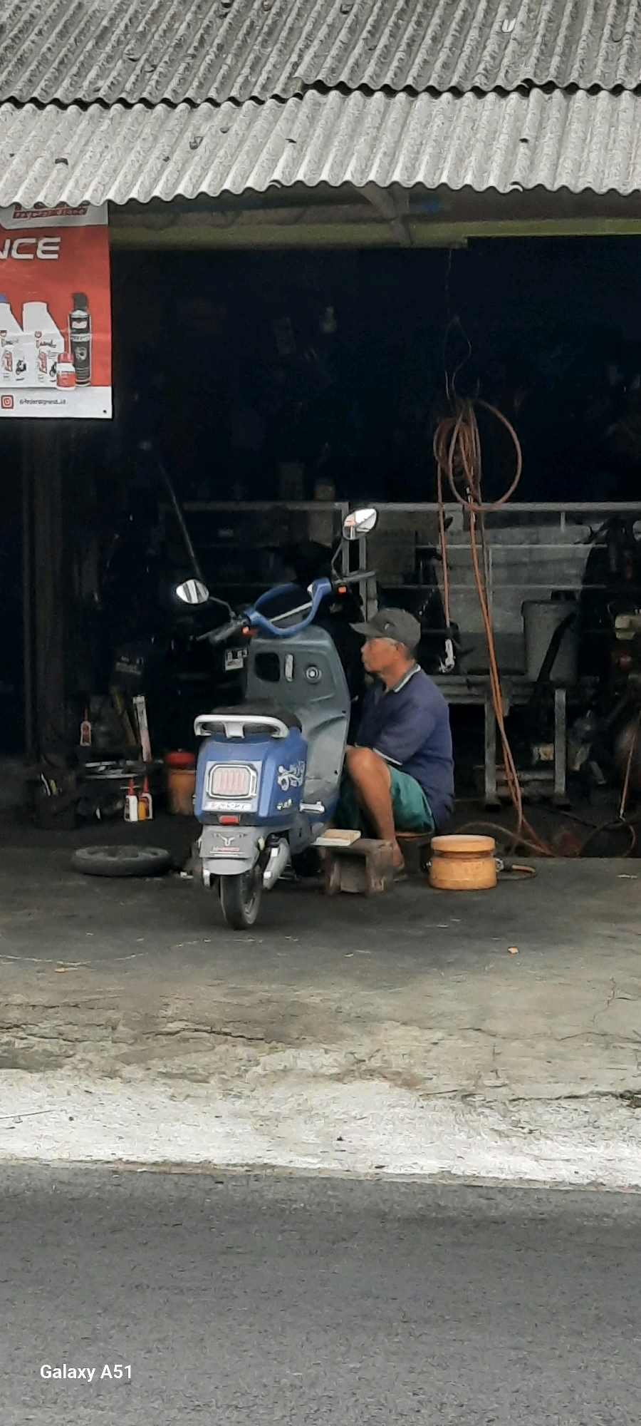 An overhead shot, likely taken from a second story, shows a man sitting on a stool next to a blue scooter inside what appears to be a repair shop in Selopuro, Indonesia. The man, wearing a gray cap, blue shirt, and green shorts, is looking to the right, away from the scooter. The scooter is parked on a gray concrete floor, with a spare tire on the ground to its left. In the background, shelves are filled with various items, and a long, coiled orange hose hangs from the ceiling. A corrugated metal roof covers the shop. A poster featuring bottles and a spray can is visible on the left side of the image, with the word "NCE" partially visible. A gray paved road runs in front of the shop. The image is tagged with "Galaxy A51".