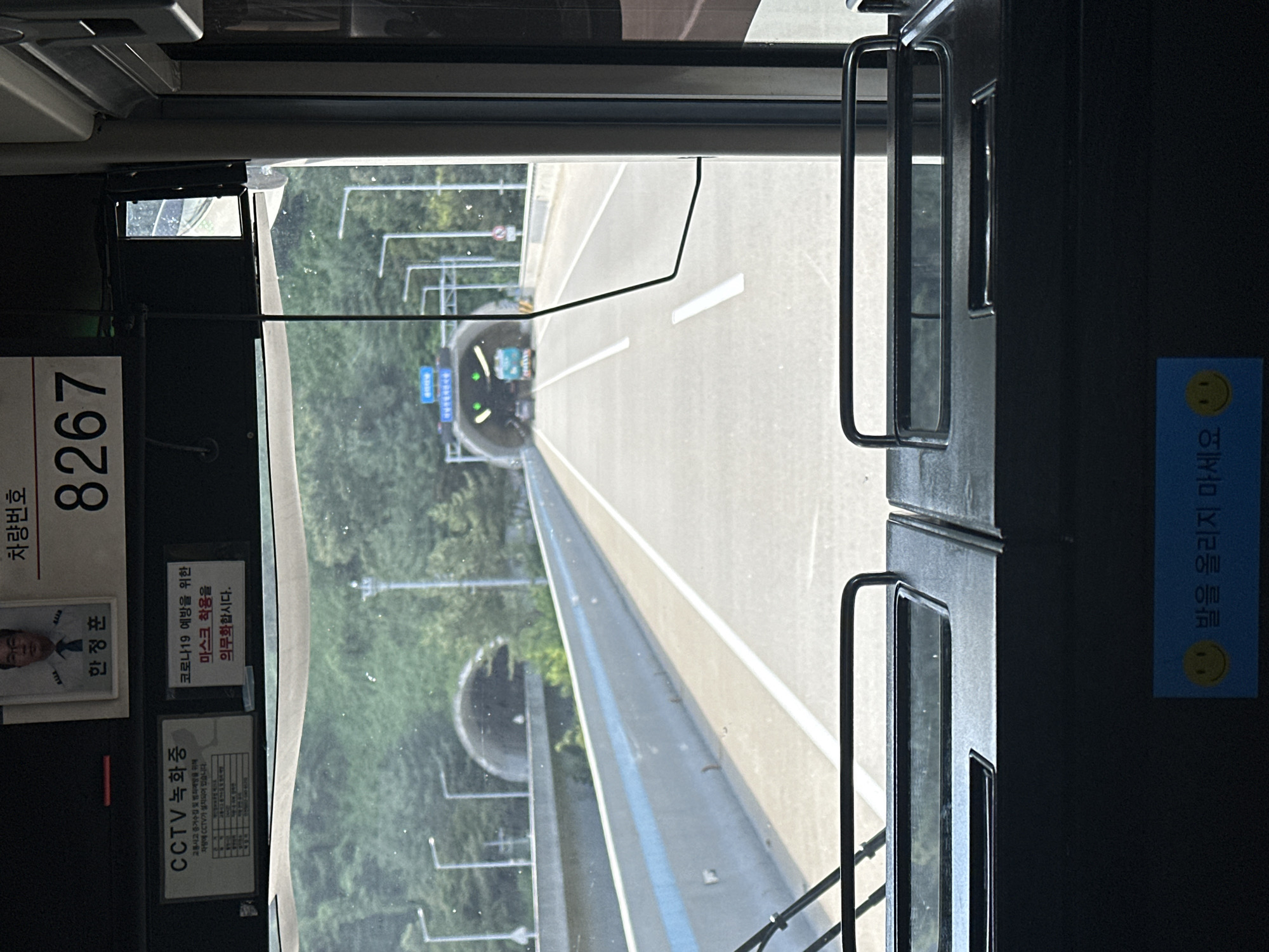 The image shows the interior of a bus, looking out the windshield. The bus appears to be traveling on a road that leads into a tunnel. A sign above the tunnel entrance is visible, displaying Korean text and green arrows indicating the direction to proceed. The road is divided by white lines and bordered by concrete barriers. Trees and a cloudy sky are visible on either side of the road.

Inside the bus, a sign with "CCTV 녹화중" (CCTV Recording) is displayed on the left. To the right of this, another sign reads "코로나19 예방을 위한 마스크 착용을 의무화합니다" (Mandatory mask wearing to prevent COVID-19). Above these signs, a photograph of a man in a uniform is present, with Korean text below him reading "한정훈". To the right of this, the number "8267" is displayed prominently. Near the bottom of the windshield, a blue sticker with a smiley face and Korean text that translates to "Don't put your feet up" is visible. The foreground shows the dashboard and seating area of the bus.

The presence of the bus, the road infrastructure, and the Korean language on the signs indicate this scene is likely in South Korea. While a specific location within Machi-ri is not provided by the image itself, the overall context suggests a transit setting on a highway.