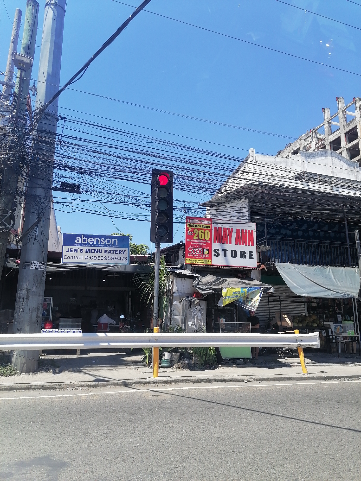The image shows a street scene in Davao City, Philippines, under a clear blue sky. A traffic light with a red signal is prominently displayed. Adjacent to the traffic light, a sign for "abenson JEN'S MENU EATERY" is visible, along with a contact number. Another sign advertises "MAY ANN STORE" with a promotion for "Stay for 2 Hours 260".
A white metal guardrail runs along the sidewalk, separating it from the road. The sidewalk is lined with various establishments, including what appears to be a store or eatery with awnings. Some people are visible inside the establishments, and a motorcycle is parked nearby. In the background, a building under construction or renovation is partially visible. Overhead, a complex network of electrical wires crisscrosses the sky. The overall environment suggests a busy urban area with commercial activity.