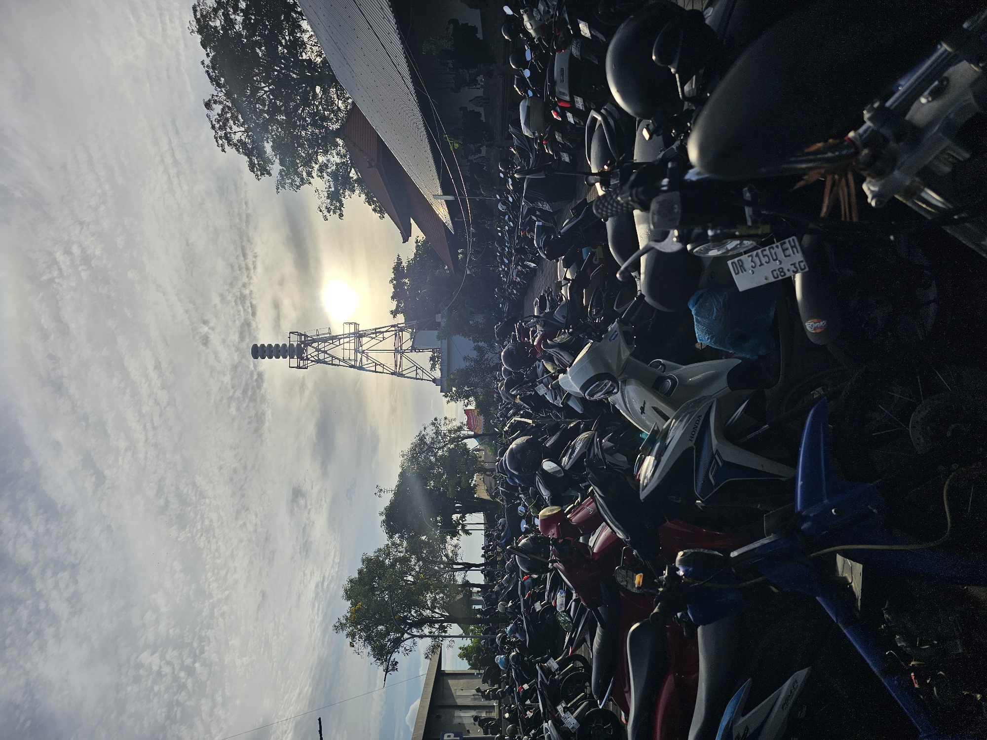 The image shows a large parking lot densely filled with motorcycles in Denpasar, Indonesia. A communications tower stands near the parking area, silhouetted against a bright, partly cloudy sky with the sun visible. The motorcycles are parked closely together, creating a crowded and tight space. Several trees line the parking area, their branches reaching towards the sky. A license plate is visible on one of the motorcycles, reading "DK 3150 EH." The image appears to be taken during daylight hours, potentially early morning or late afternoon, given the angle of the sun.