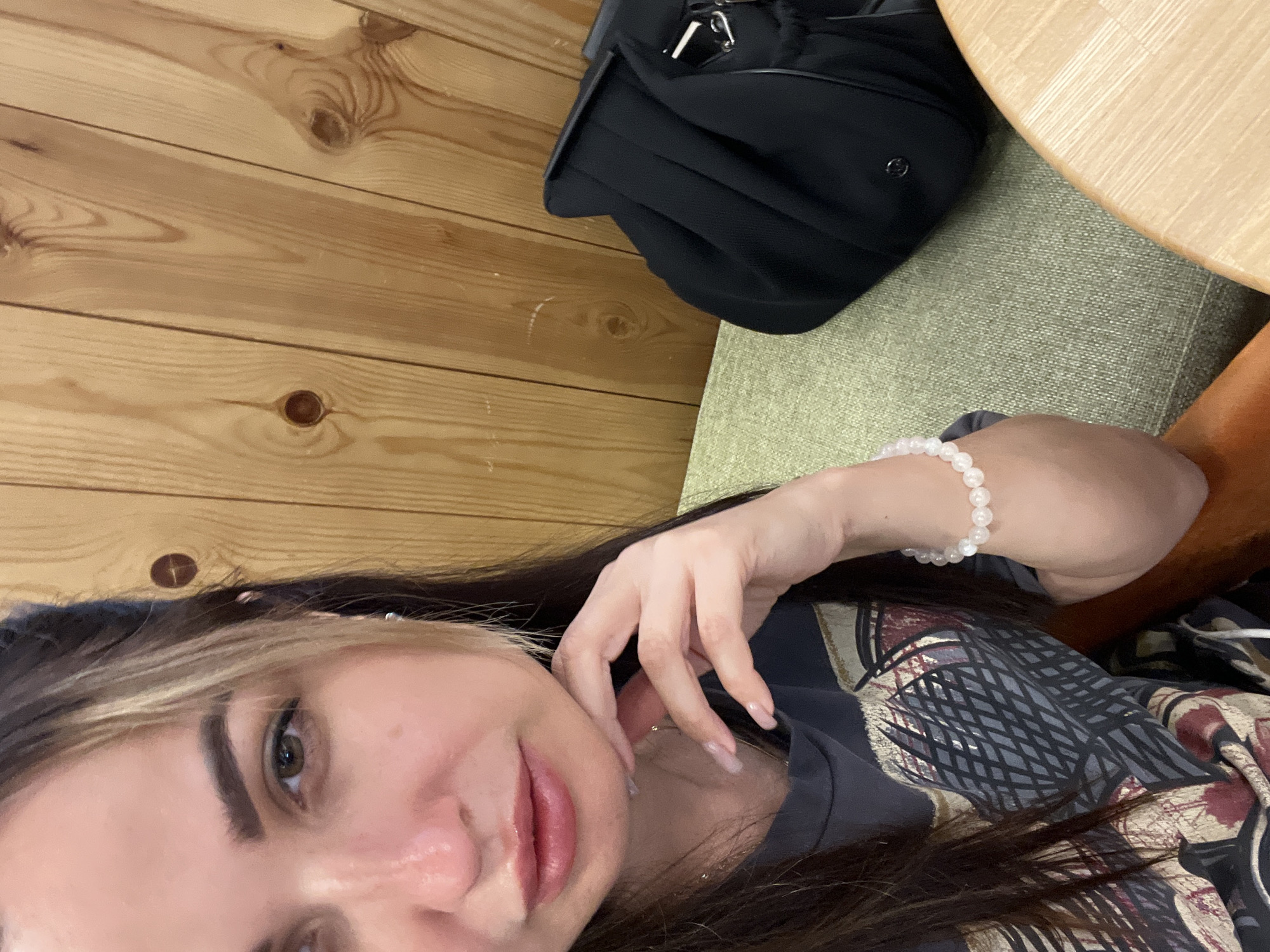 A woman is captured in a close-up, over-the-shoulder selfie, her face angled towards the camera. She has brown hair with blonde highlights, framing her face. Her left hand rests on her chin, and she wears a bracelet of white beads on her right wrist. She's dressed in a dark t-shirt with a graphic print. Behind her, a black bag is visible on a textured green seat, against a backdrop of light brown wooden paneling. The setting appears to be indoors, possibly a cafe or a restaurant, with a round wooden table partially visible in the foreground. The lighting is soft, and the overall atmosphere is casual and relaxed.