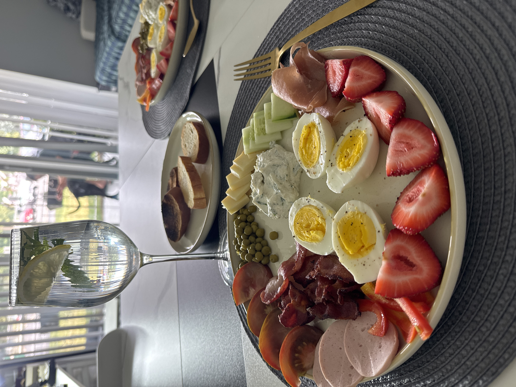 A close-up shot showcases a delectable breakfast spread artfully arranged on a plate. The plate is laden with an assortment of food items, including sliced hard-boiled eggs, crispy bacon strips, thinly sliced bologna, plump peas, and halved strawberries. Accompanying these are slices of tomato, cucumber, red and yellow bell peppers, and cheese cubes. A dollop of creamy dip, possibly cream cheese with herbs, sits at the center, with a few slices of toasted bread in a separate small bowl visible in the background. A glass filled with water, lemon slices, and mint leaves stands prominently to the left, catching the light. A golden fork rests on the right side of the plate. The setting appears to be an indoor dining area, possibly a home, with a modern white table and a window in the background revealing a blurred outdoor scene. The overall impression is one of a well-prepared and visually appealing breakfast. There is no information about the location in the city of Wheeling, United States of America.