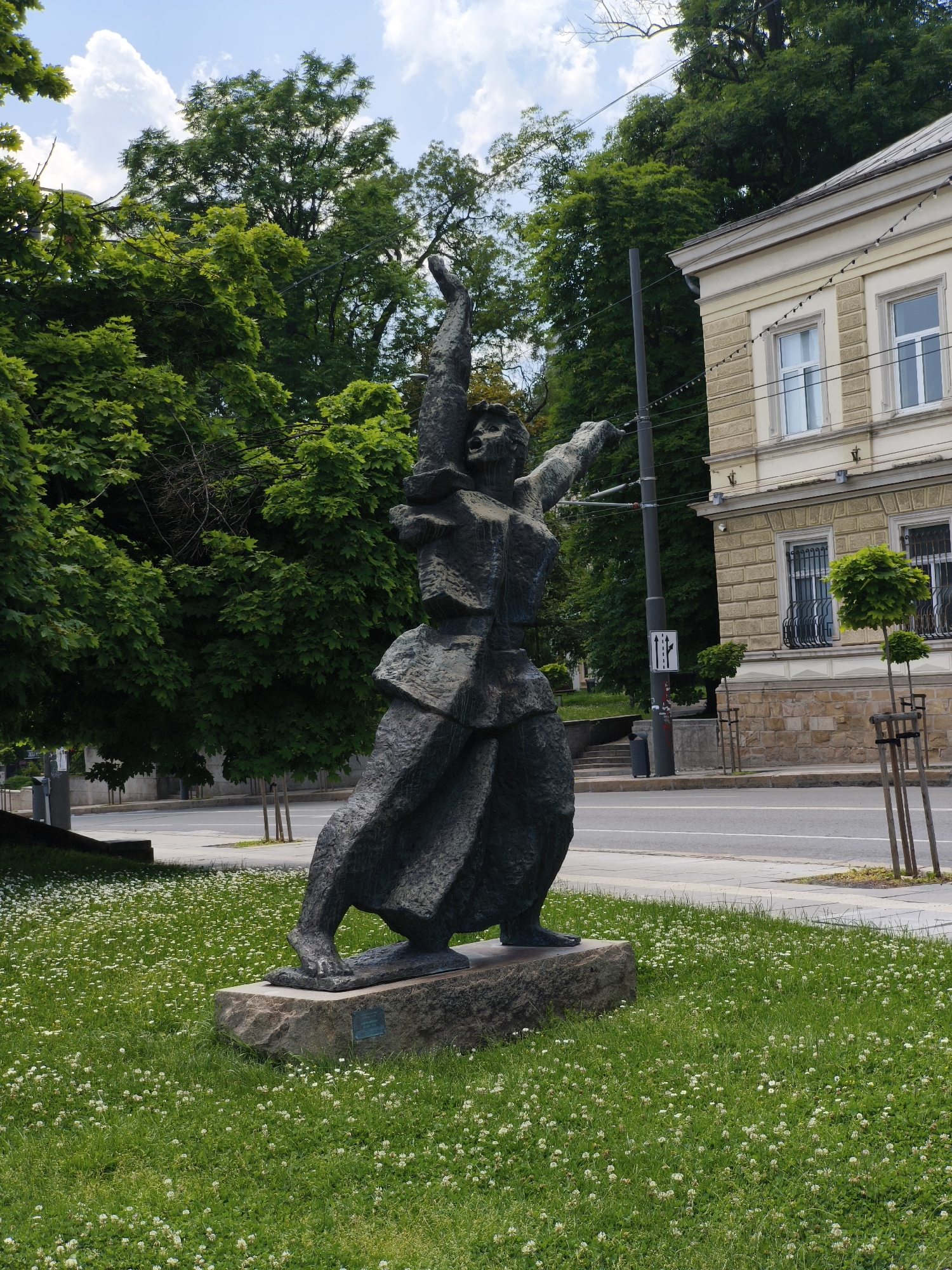 The image displays a detailed view of the "Monument to the Bulgarian Revolutionaries" (also known as "The Call" or "The Uprising") by Georgi Chapkanov, erected in 1976, located in Sofia, Bulgaria, specifically on the green strip along Tsar Osvoboditel Boulevard, near the intersection with Georgi S. Rakovski Street, in proximity to the Doctor's Garden.
The primary subject is a dark-patinated bronze or metal sculpture of a human figure, rendered in a dynamic, expressive, and somewhat fragmented style. The figure appears to be stepping forward or ascending, with its left arm raised high, holding an indistinct object, and its mouth open as if in a shout or call. The right arm is extended outwards to the right. The sculpture rests on a rough-hewn stone plinth, on which a small identification plaque is affixed.
The sculpture is positioned on a vibrant green lawn densely populated with small white clover flowers. Behind the lawn, a grey concrete sidewalk borders an asphalt road with two visible lanes. To the immediate left of the sculpture, a dense mass of deciduous trees with bright green foliage fills the midground, with branches extending into the upper left of the frame. Above the trees, a bright blue sky with scattered white cumulus clouds is visible.
To the right of the sculpture and across the road, a multi-story building with a light-colored facade is present. The building exhibits classical architectural elements, including a defined cornice, rectangular windows with white frames, and decorative black iron grilles on the lower-level windows. The ground floor features a rusticated stone texture, while the upper floor appears to be smooth plaster. Several strings of small, white decorative lights are strung horizontally across the building's facade. In front of the building, a row of slender, newly planted trees with wooden support stakes lines the sidewalk. A tall, dark grey utility pole with multiple overhead wires stands between the sculpture and the building. Attached to this pole is a white rectangular traffic sign depicting three vertical arrows: two pointing straight ahead and one pointing to the left. A grey waste receptacle is partially visible near the base of the utility pole. The scene is illuminated by clear daylight, casting minimal shadows directly observable.