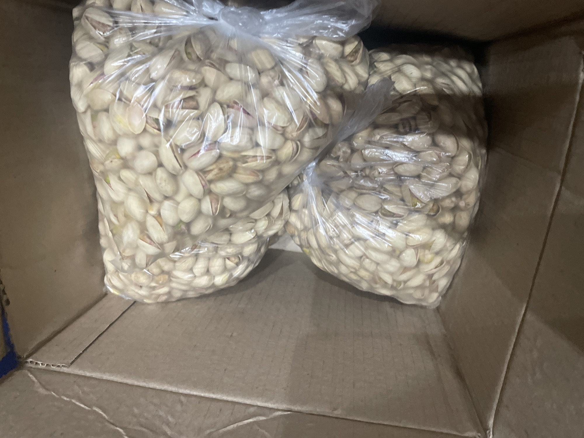 The image shows two transparent plastic bags filled with pistachios inside a cardboard box. The box is open, and the bags of pistachios are stacked on top of each other. The pistachios are light beige and green, with some shells open. The cardboard box is brown and appears to be made of corrugated cardboard. The lighting is dim, casting shadows on the pistachios and the inside of the box. The scene is indoors, likely in a storage area or warehouse. There are no people or animals visible in the image. The image is taken from a slightly elevated angle, looking down into the box. The specific location in the city of Zhukovo, Russia, is not discernible from the image itself.