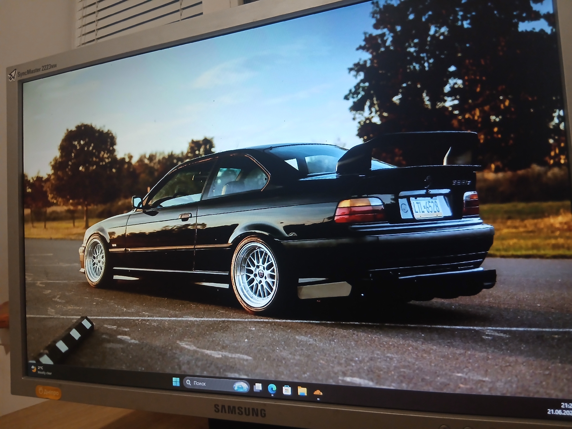 The image displays a computer monitor showing a wallpaper of a black BMW M3 E36 coupe parked on an asphalt surface. The car has a prominent rear spoiler and custom wheels. In the background, there are trees with autumn foliage and a clear blue sky. The monitor itself is a Samsung SyncMaster 2223NW. The lower part of the screen shows the Windows taskbar with icons and a search bar. A circular orange sticker with Cyrillic text, likely indicating the city of Dnipro, Ukraine, is visible on the lower right side of the monitor bezel. A small clock displaying "21:2", and "21.06.2023" is present on the taskbar. There is no indication of the car's location being in the city of Dnipro, Ukraine, other than the sticker on the monitor.