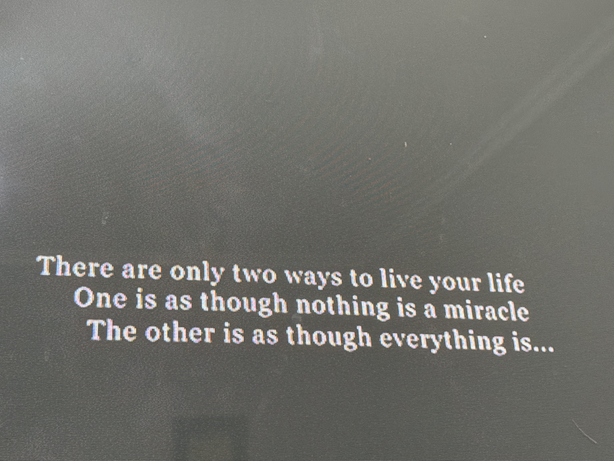 The image displays a philosophical quote presented in white serif font against a dark grey, textured background, characteristic of a digital screen or monitor. Fine horizontal lines and subtle pixelation are visible across the background and within the letterforms, indicating a digital display. The text reads: "There are only two ways to live your life One is as though nothing is a miracle The other is as though everything is..." The quote ends with an ellipsis, suggesting an incomplete thought. There are no people or distinct objects in the frame, and no specific activity is taking place beyond the static display of the text. The overall scene conveys a reflective mood due to the nature of the quote. Based on the provided context, the image was taken in Tbilisi, Georgia.