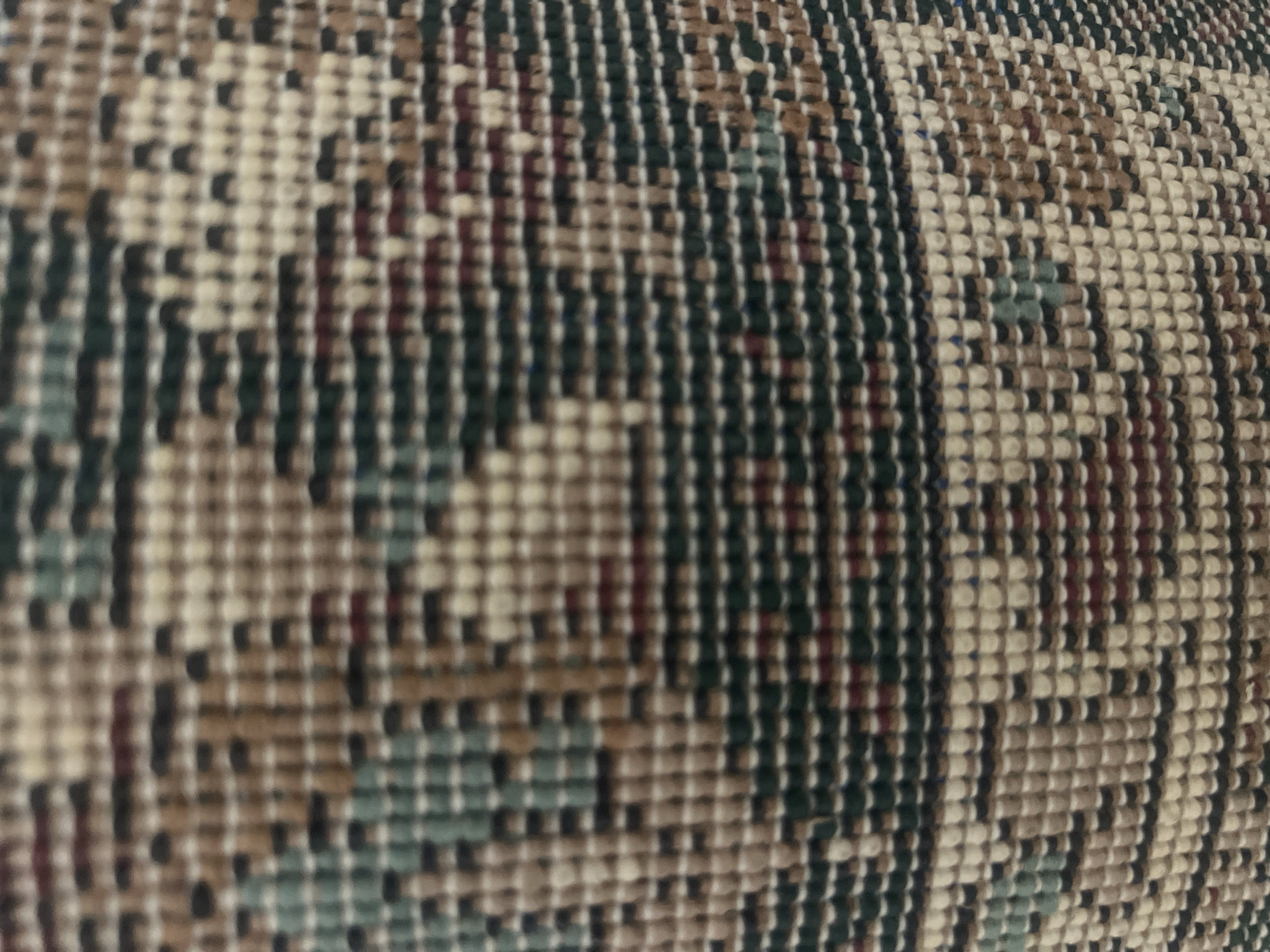 The image displays a close-up, high-magnification view of a woven textile, likely a rug, carpet, or upholstery fabric. The texture is dense and appears to be formed by numerous individual, short, cylindrical or squared-off thread tufts arranged in a grid-like structure, giving the pattern a distinctively pixelated or blocky appearance.
The pattern is intricate and geometric, composed of repeating abstract motifs. These motifs include stepped lines, chevron-like shapes, and larger block formations, suggesting a traditional or ethnic design. The colors employed are muted and earthy, forming a complex palette:
- Dominant colors include various shades of beige and cream, appearing as the lightest elements.
- Darker tones consist of deep forest green or dark teal, dark brown, and black.
- Accent colors include a reddish-brown or maroon, and faint hints of light blue or grey are present in some areas.
- The arrangement of these colored tufts creates distinct boundaries and transitions within the pattern.
The photograph exhibits a soft focus, rendering the finer details slightly blurred while still clearly defining the overall pattern and color distribution. There are no discernible objects, individuals, actions, or environmental cues within the frame that allow for the determination of a specific location in Casablanca, Morocco, or any other geographic area. The scene is solely a detailed macro shot of the textile's surface.