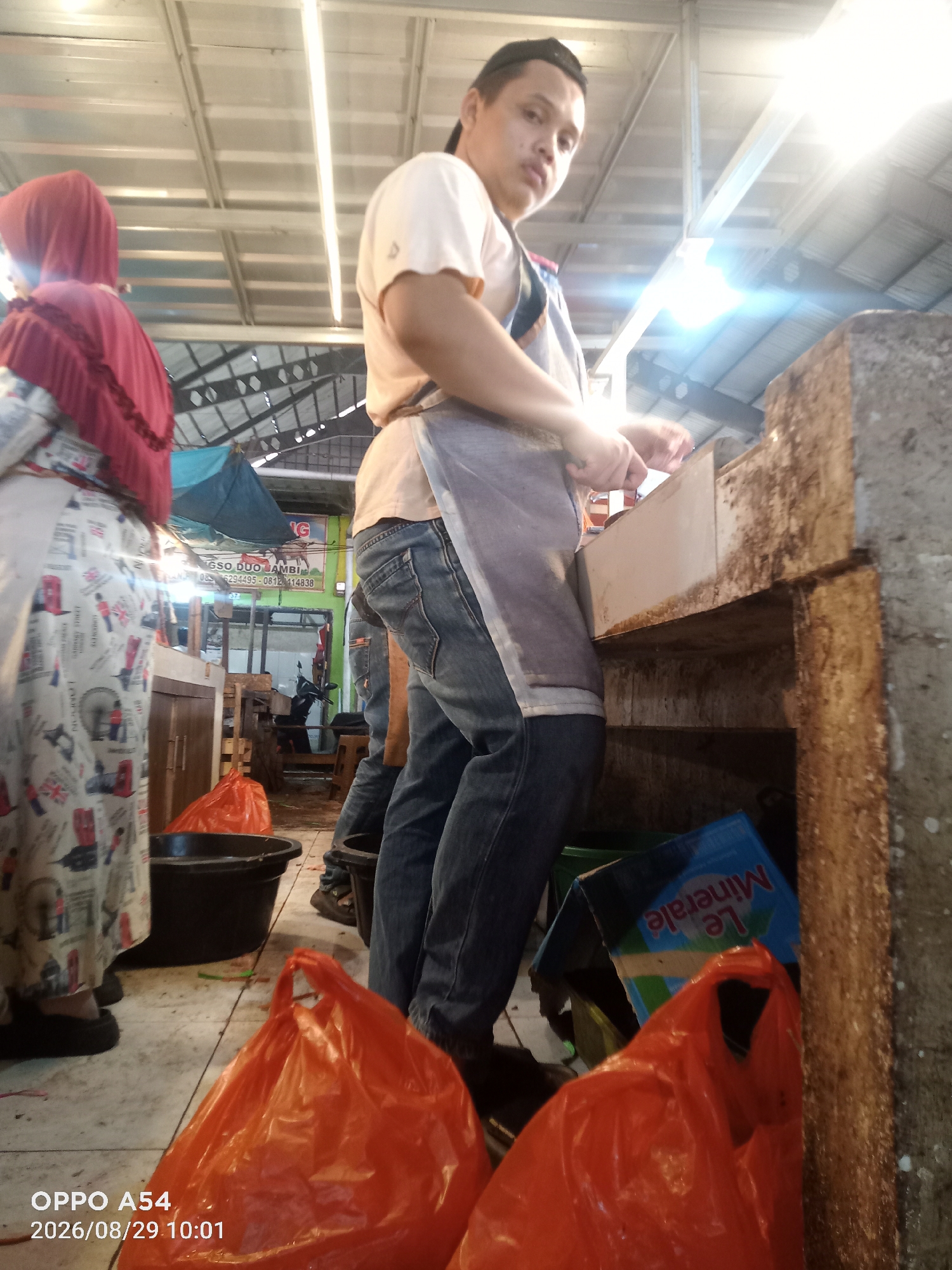 The image depicts a busy market scene in Jambi, Indonesia. In the foreground, a man wearing a light-colored t-shirt and jeans is seen preparing something at a counter. He is partially obscured by an apron. To the left, a woman wearing a red hijab and a patterned dress is visible. Two bright orange plastic bags are prominent in the lower portion of the image, suggesting recent purchases or items for sale. Black buckets are also present, one containing what appears to be a blue box with text, possibly "Le Minerale". The environment appears to be an indoor market with a corrugated metal ceiling and bright lighting. The overall impression is one of a functional, everyday market setting.