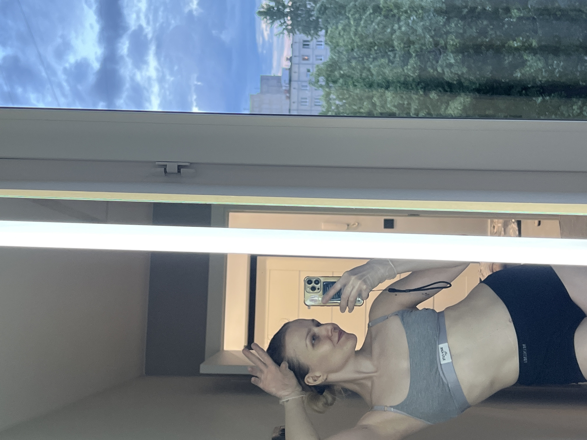 A reflection of a woman in workout attire is captured in a mirror. The woman is wearing a gray sports bra and black shorts. She is holding a cell phone and taking a selfie. Outside the window, there is a view of apartment buildings and trees, with a cloudy sky. The location appears to be Khimki, Russia, based on the style of architecture and urban environment visible through the window.