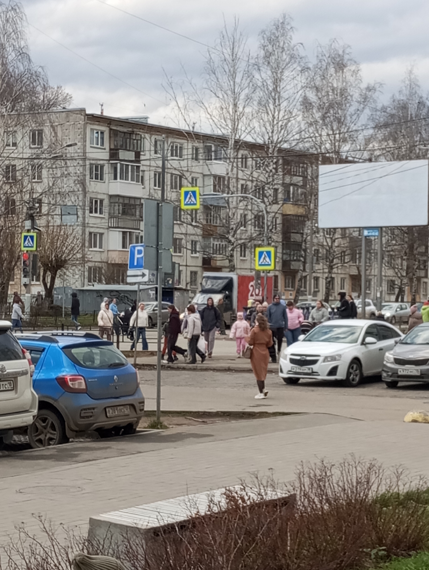 The image captures a bustling street scene in an urban, residential area under an overcast sky, suggesting daytime in a cold season, possibly late winter or early spring, given the bare trees. Numerous pedestrians, dressed in winter-appropriate clothing like jackets and coats, are seen crossing a marked pedestrian crossing.
In the foreground, a white SUV is partially visible on the left, next to a blue Renault Duster parked with its rear facing the viewer, displaying a partially visible license plate "E 281 OK 18". On the right, a concrete bench and some dormant bushes are present.
The mid-ground is dominated by the pedestrian crossing, where a crowd of people is gathered. A woman in a long, light brown coat walks towards the left. Others include individuals pushing strollers and a child in a pink outfit. Cars are stopped on the road, seemingly waiting for pedestrians. A white sedan, possibly a Chevrolet Cruze, has a partially visible license plate "P 192 PP 193," and a grey sedan shows "X 977 EC 193."
The background features multi-story apartment buildings, characteristic of post-Soviet architecture. Bare trees with intricate branches are scattered throughout. Several street signs are visible: three yellow square pedestrian crossing signs, a blue square parking sign with a white 'P' and a right-pointing arrow, and a traffic light showing a red signal. A large red sign with the number "20" is visible on a building or poster in the distance. Overhead wires crisscross the sky above the buildings. The scene depicts everyday activity in a busy city environment.