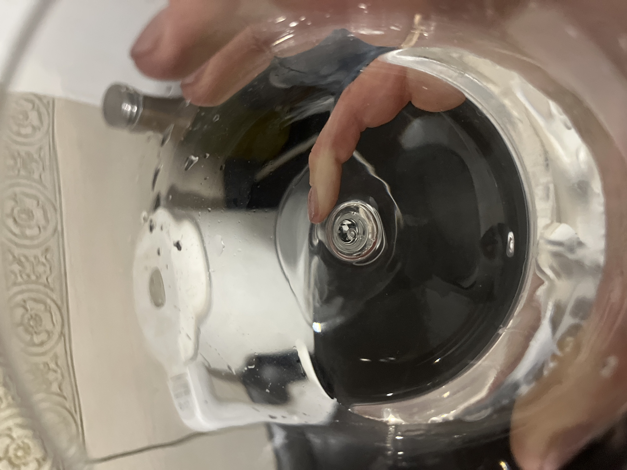 The image shows a close-up, interior view of a clear glass, likely a drinking glass or vase, with water inside. A person's hand is visible in the foreground, with fingers reaching into the glass. The hand appears to be interacting with the water or an object submerged within it. A small, metallic object, possibly a cap or a lid, is visible submerged in the water, with a finger touching it. Reflections and distortions are present due to the water and the curved glass. In the background, through the glass, a white object with decorative molding and what appears to be a silver cap or lid of another container are dimly visible. The lighting suggests an indoor environment. The image does not contain any specific information that would allow for its location to be identified in the city of Budva, Montenegro.