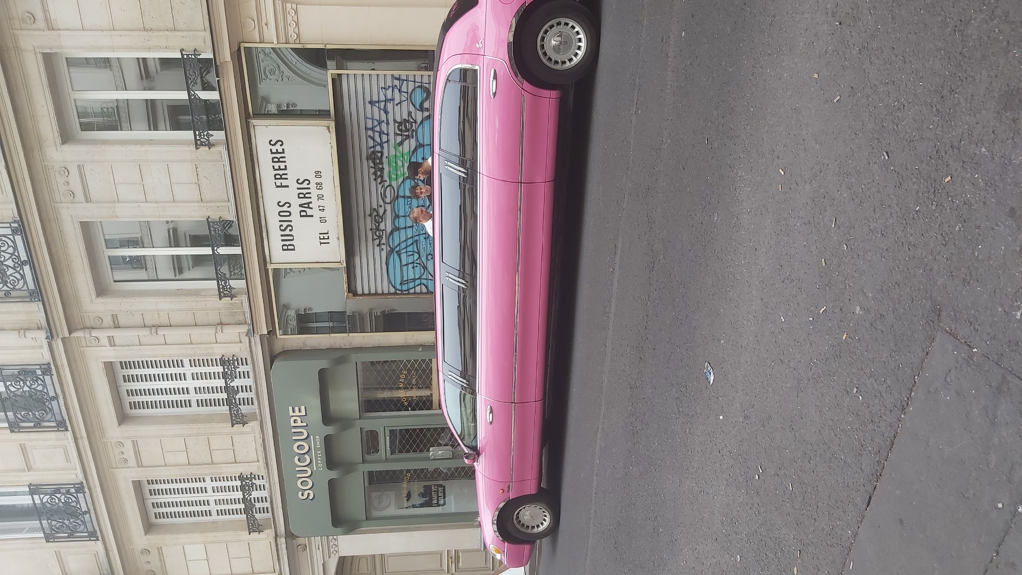 A long, pink stretch limousine is parked on a street in Paris, France. The limousine is the main subject of the image, and it occupies most of the frame. The street is paved with asphalt, and there is a sidewalk on the left side of the image. In the background, there is a building with a light-colored facade and several windows. Above a shop with the name "SOUCOUPE COFFEE SHOP" on it, a white sign reads "BUSIOS FRERES PARIS" and a phone number. Three young men are visible in the back window of the limousine, looking out. One is wearing a white shirt, and the other two are in casual clothing. The overall impression is a bright, sunny day in an urban setting.