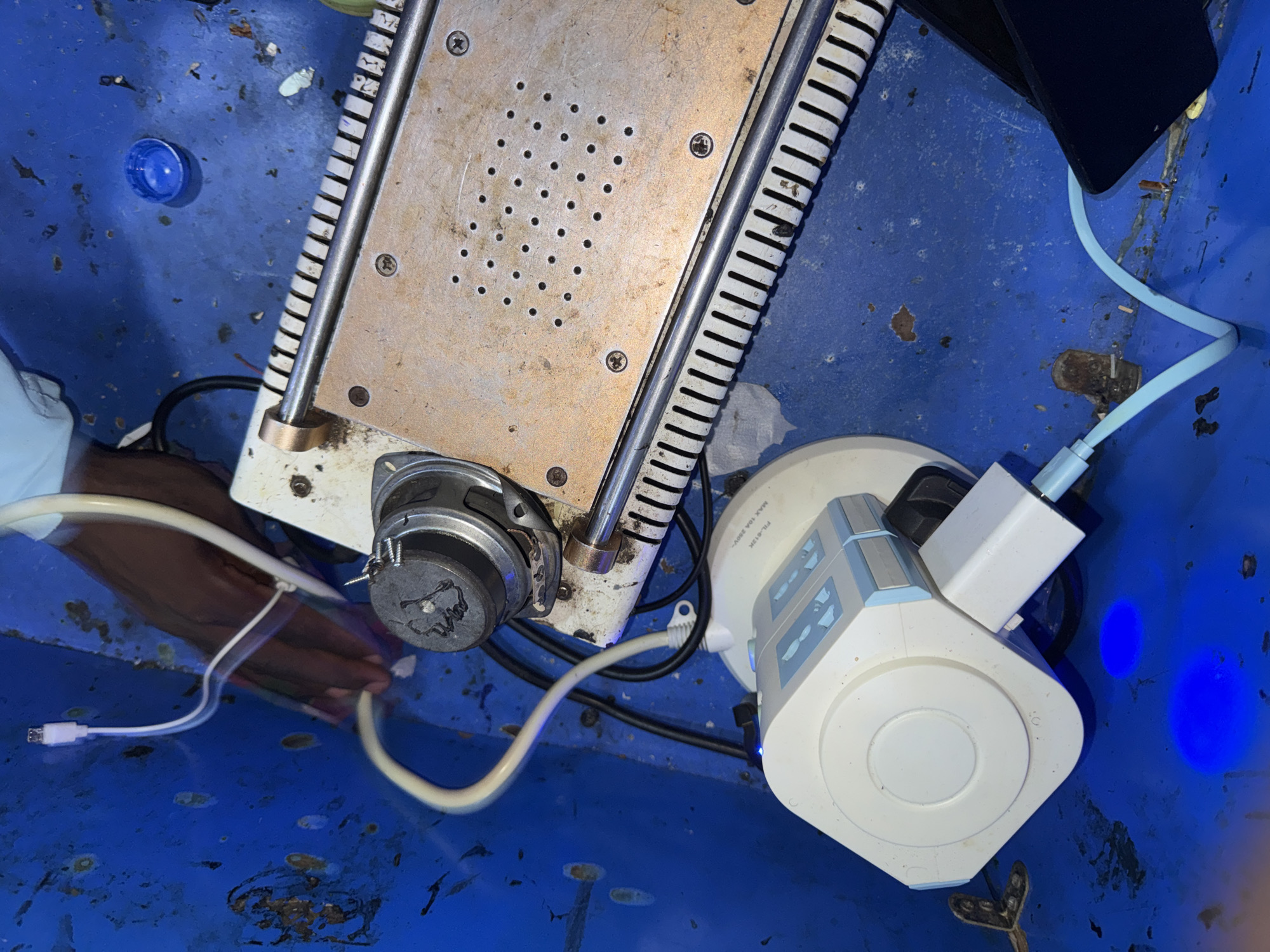 The image shows a close-up, overhead view of a blue plastic container filled with various electronic components and a blue bottle cap. In the center is a metallic object with a grid of small holes on its surface, possibly a heat sink or part of a device. To the right of this object is a white power strip with multiple sockets and a blue light glowing from within. A light blue USB cable is plugged into the power strip and runs towards the top right of the frame, where it connects to a dark object, likely a phone or tablet. Another white USB cable extends from the bottom left of the frame, leading towards the metallic object. A small, silver speaker is attached to the metallic object. The blue container appears to be dirty, with some rust spots and debris scattered on its surface. The scene suggests a workspace or a storage area for electronic equipment, possibly in Jalingo, Nigeria, given the location context. There is no clear indication of the time of day or weather. The overall impression is one of disarray and utility, with a focus on electronic devices and their connectivity.